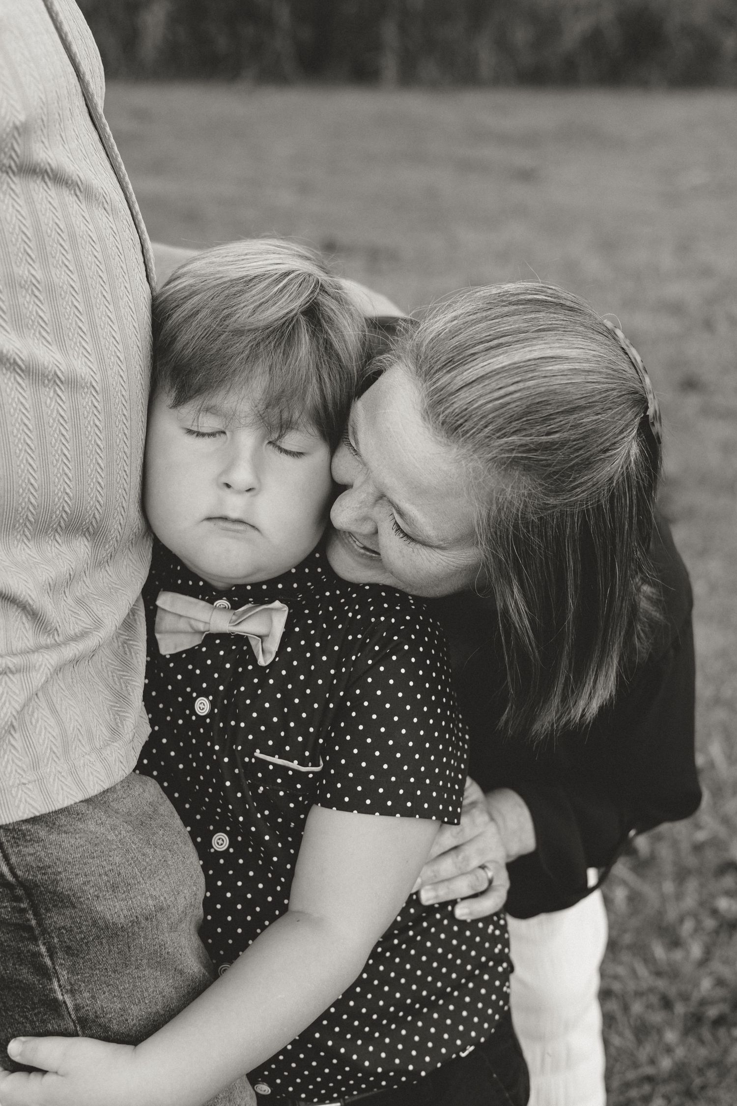 A mother tenderly kisses her young son as he leans against his father outdoors in a black and white family portrait.