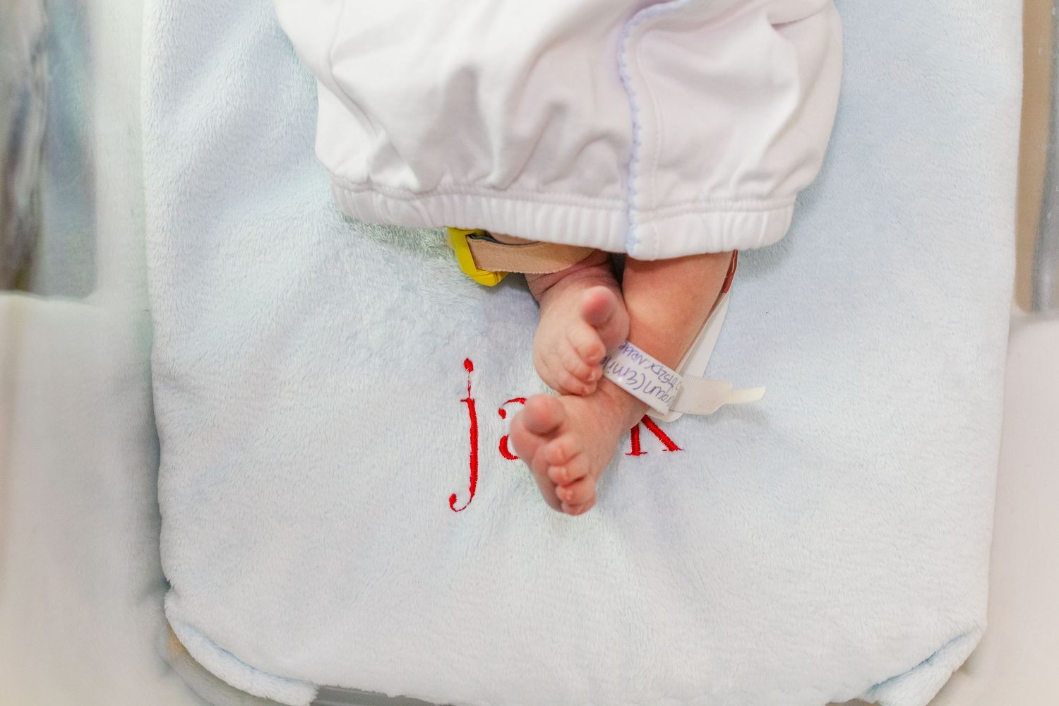 Newborn baby feet peeking from white swaddle blanket with red embroidered name in hospital bassinet.