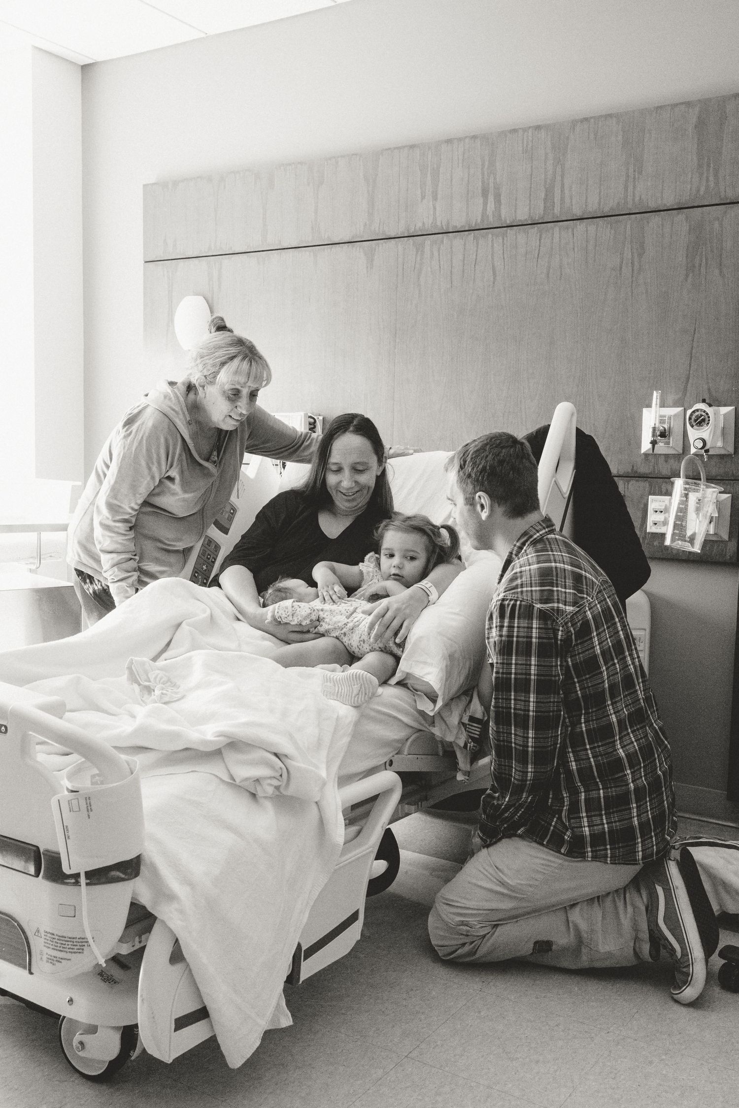 Family gathers around mother in hospital bed holding newborn and toddler.