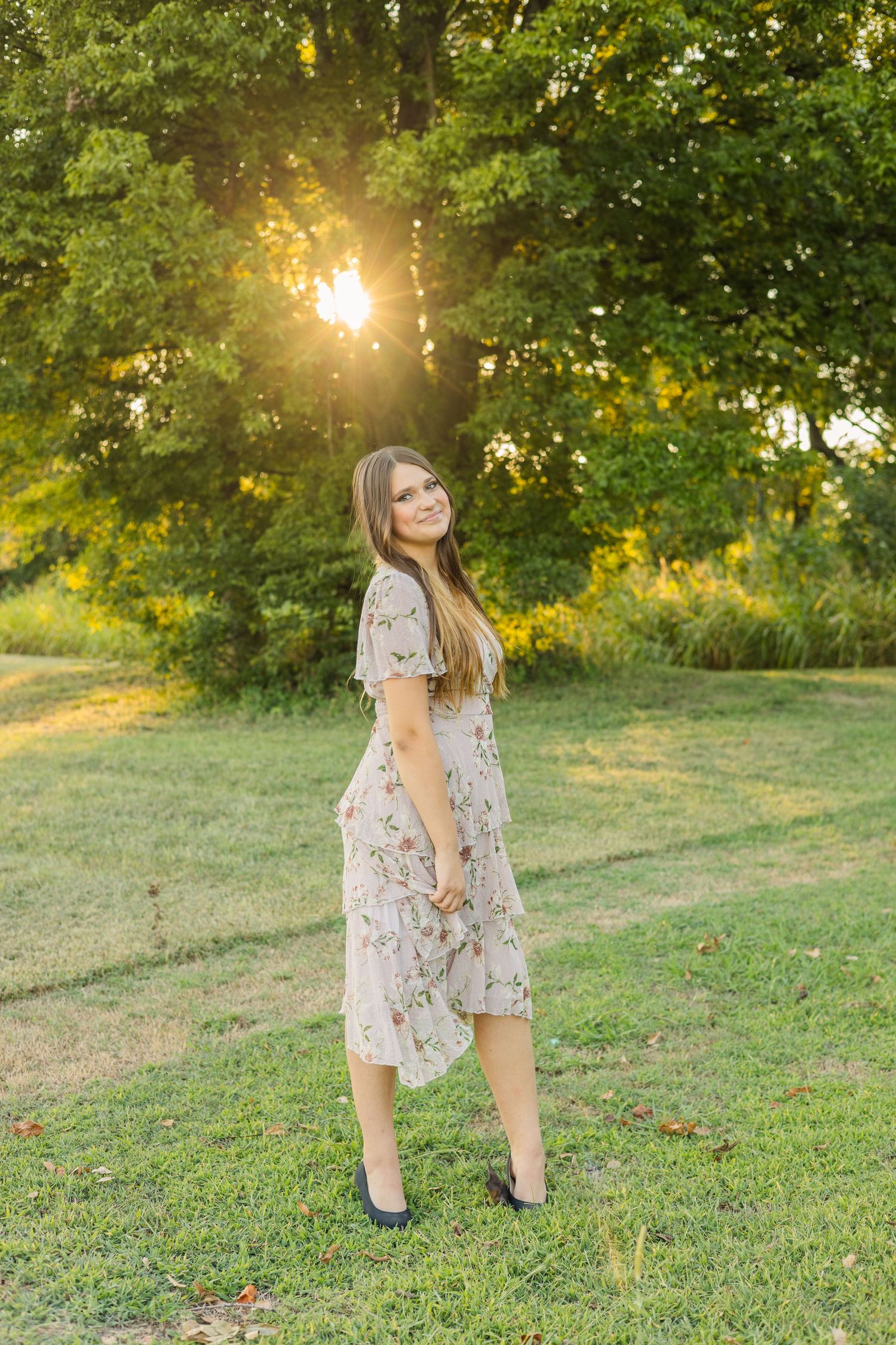 A young woman in a floral midi dress standing on green grass with golden sunset light behind her.