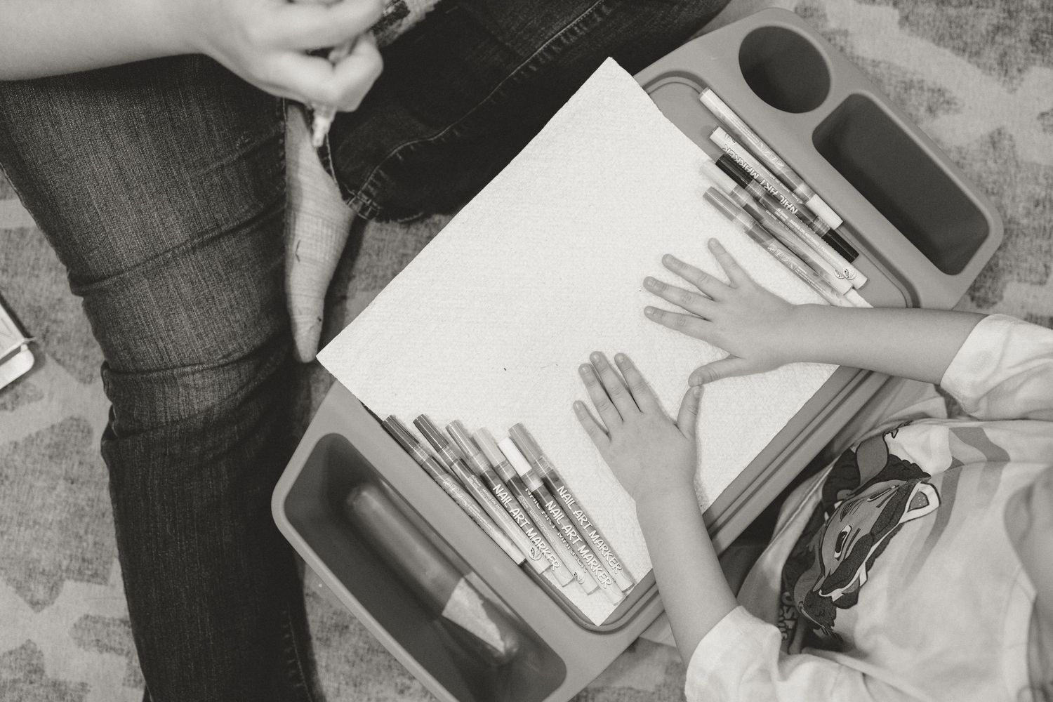 Black and white overhead view of a child painting with watercolors on white paper indoors.