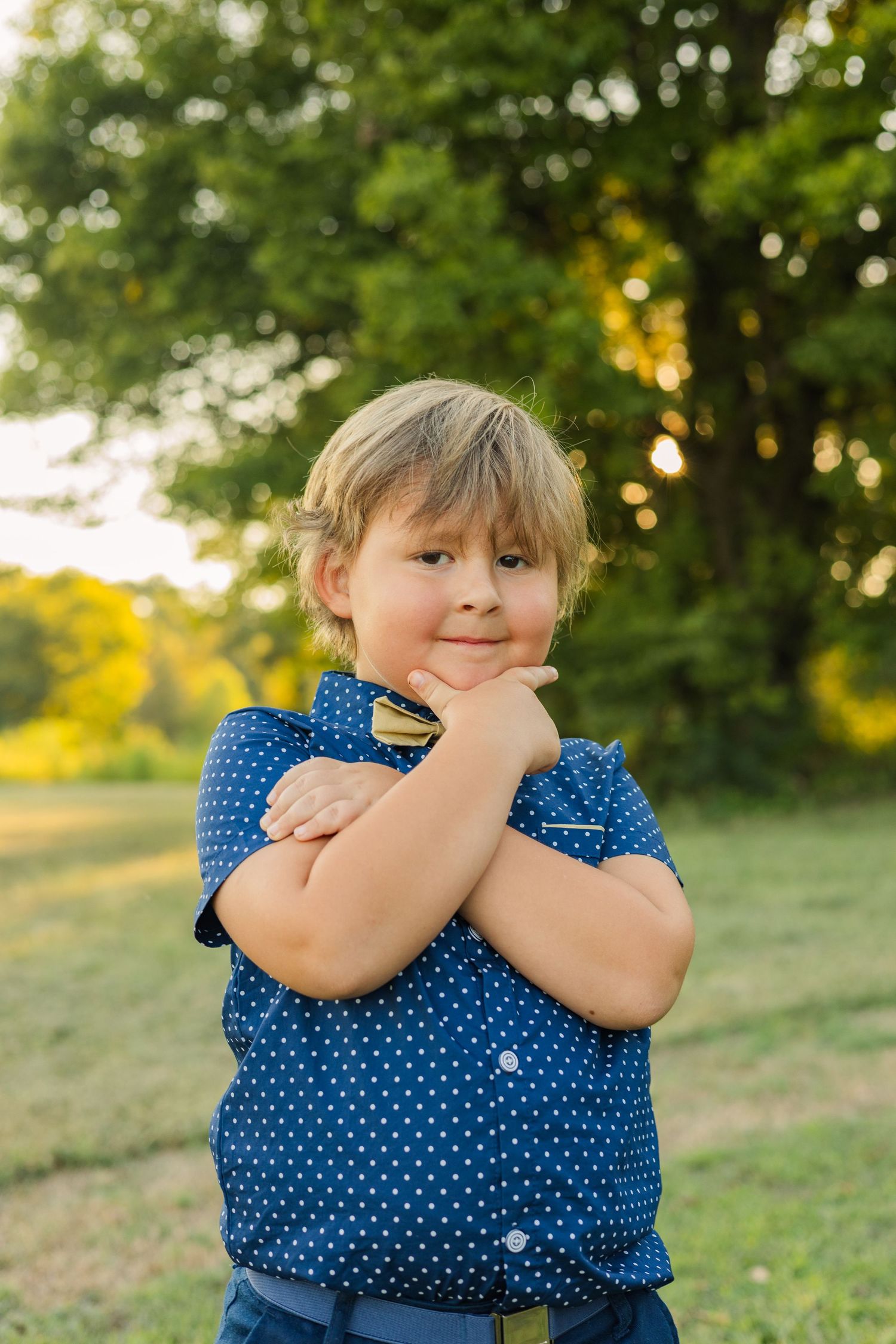 Young blonde boy in blue polka dot shirt posing confidently with arms crossed in a sunny park.
