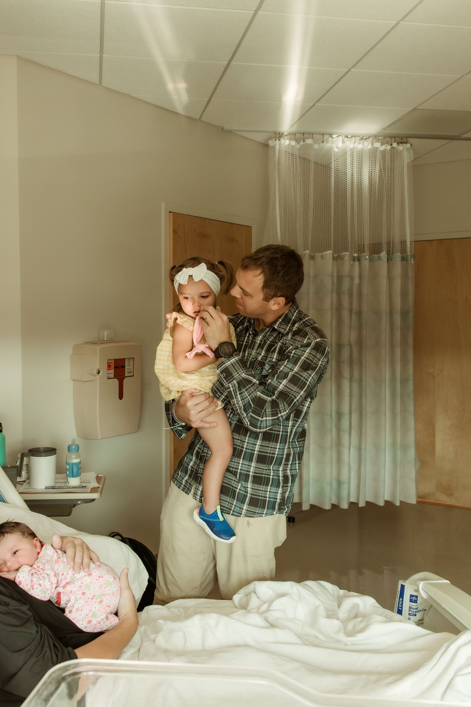 Father holds toddler girl in hospital room while mother cradles newborn baby in bed nearby.