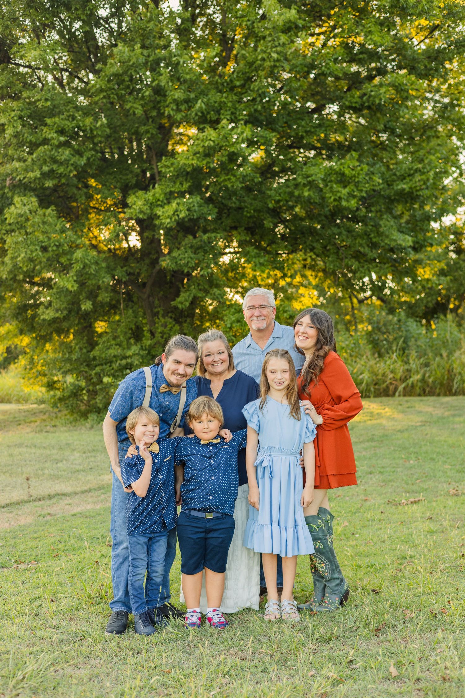 Multi-generational family portrait of seven people posing together on a green lawn with large trees behind them.