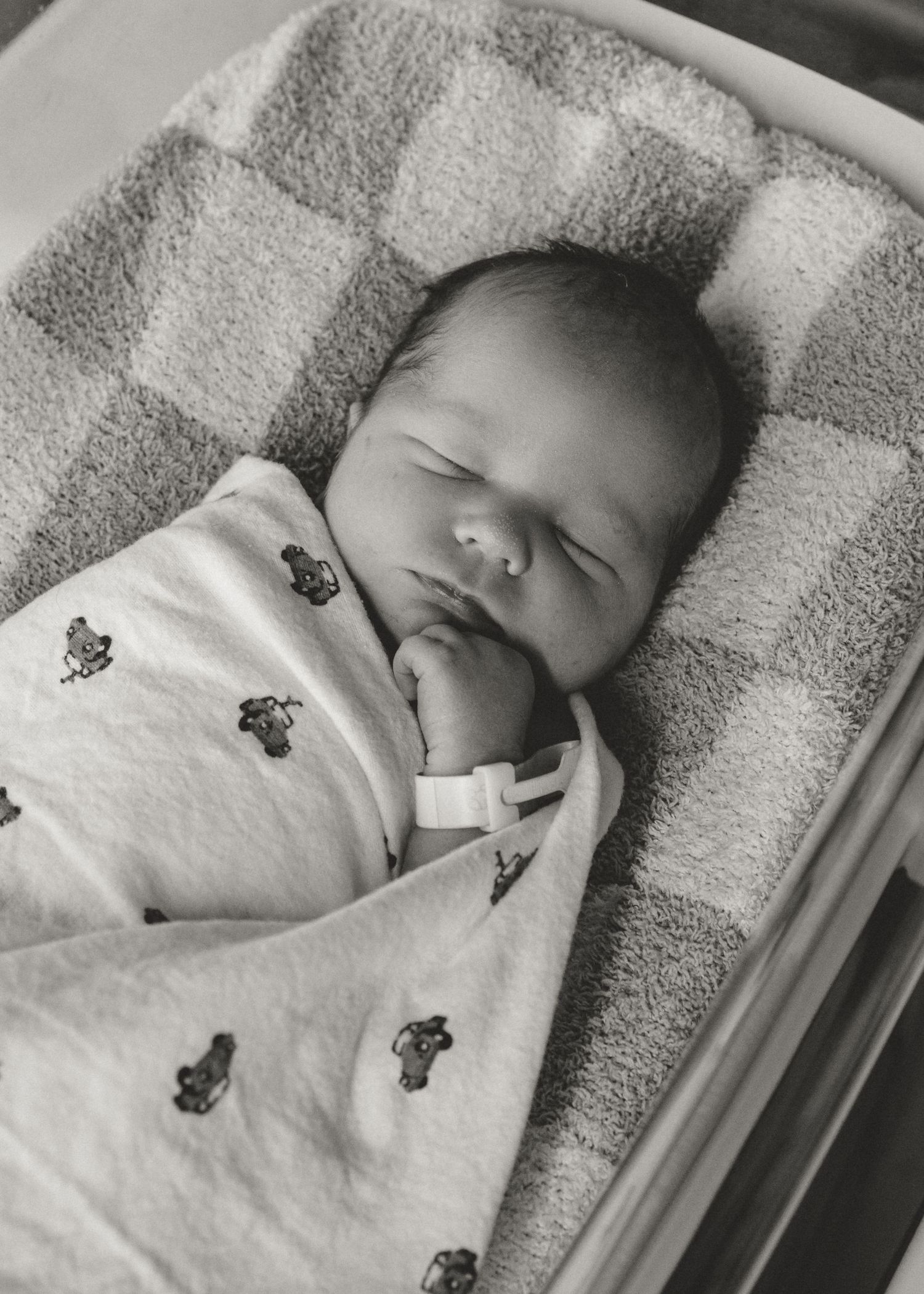 Black and white photo of a sleeping newborn baby wrapped in a patterned blanket inside a hospital bassinet.