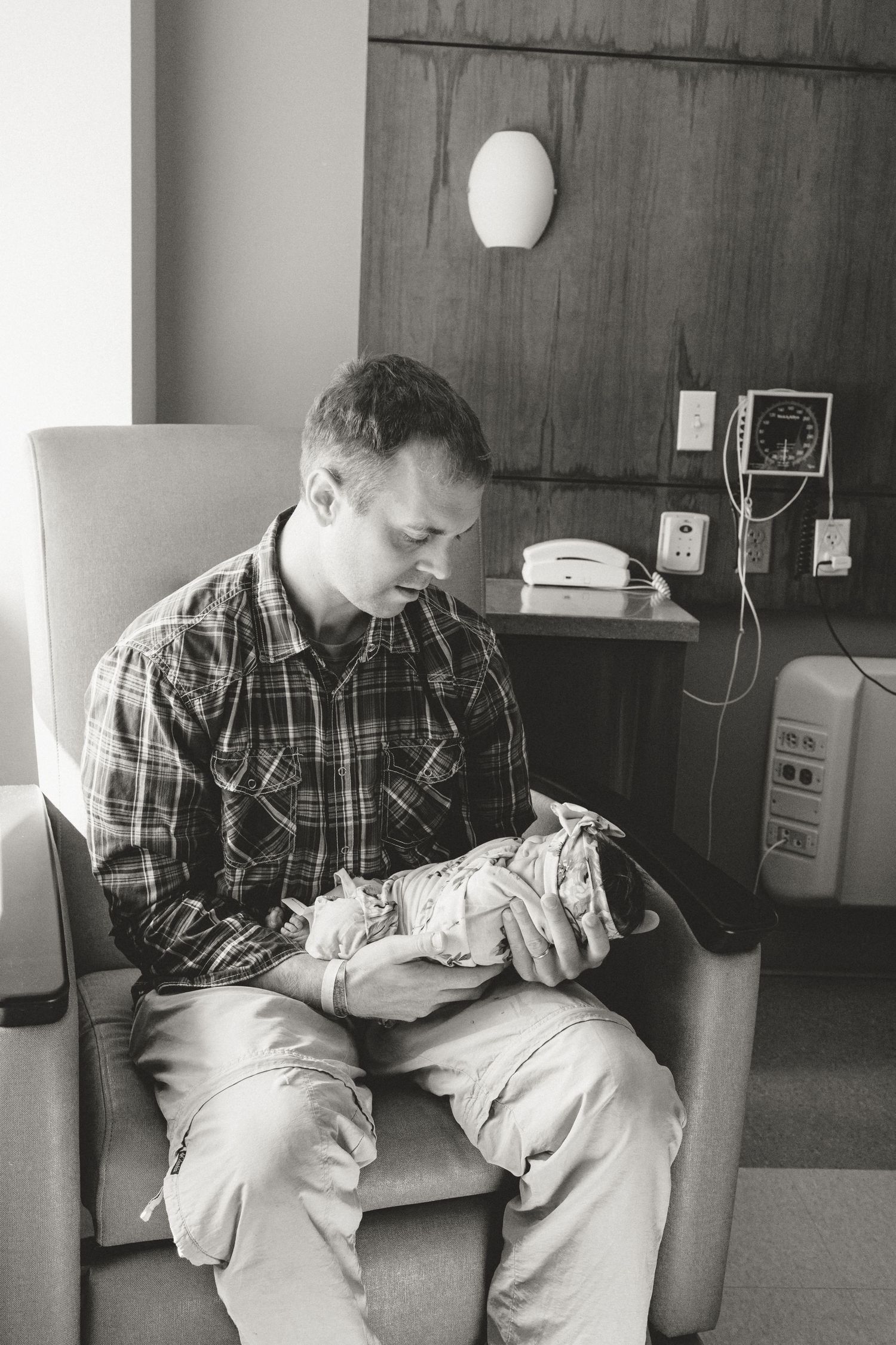 Father holding newborn baby in hospital room chair, black and white photo.