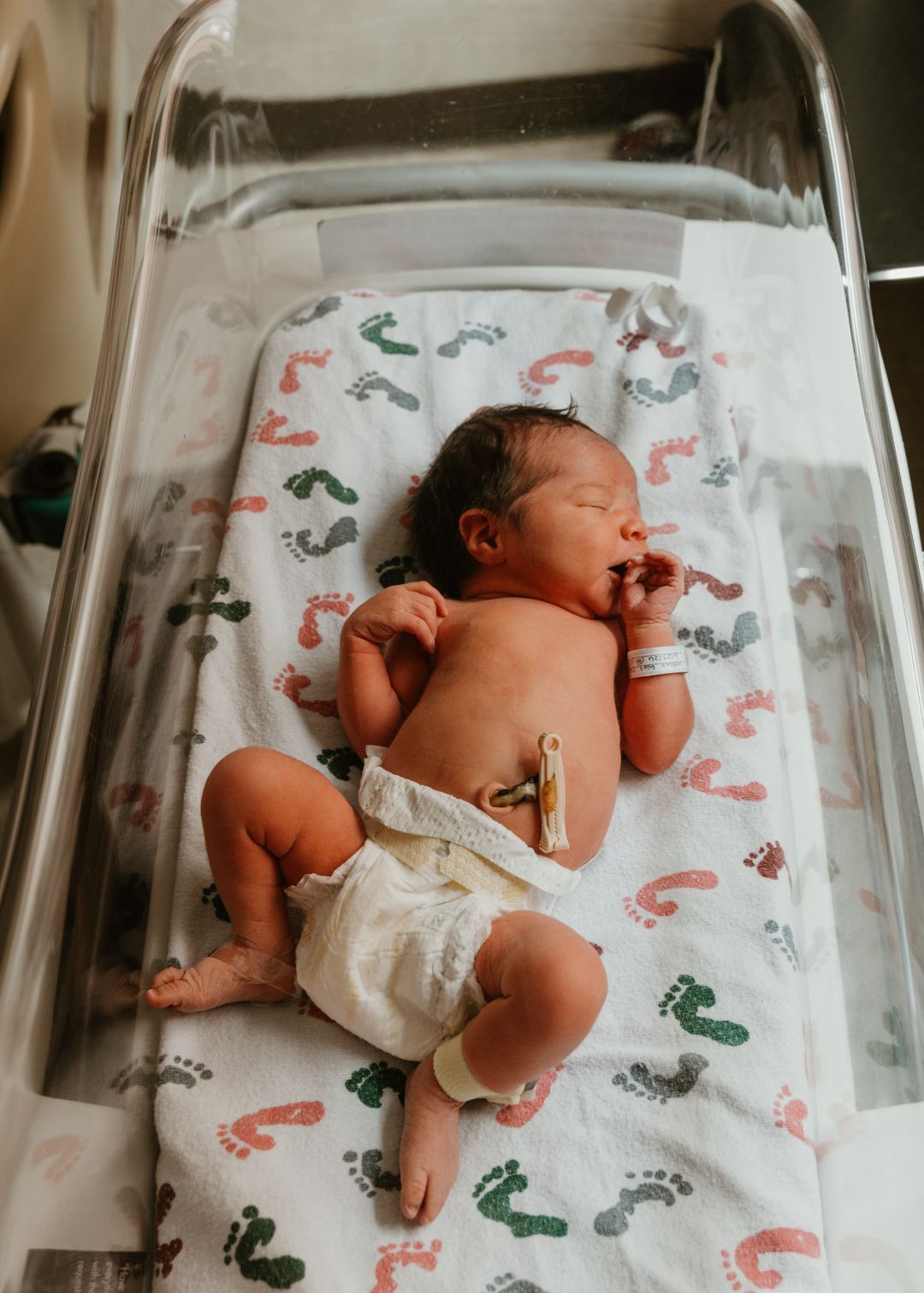 Newborn baby in diaper lying in hospital bassinet on footprint patterned blanket.