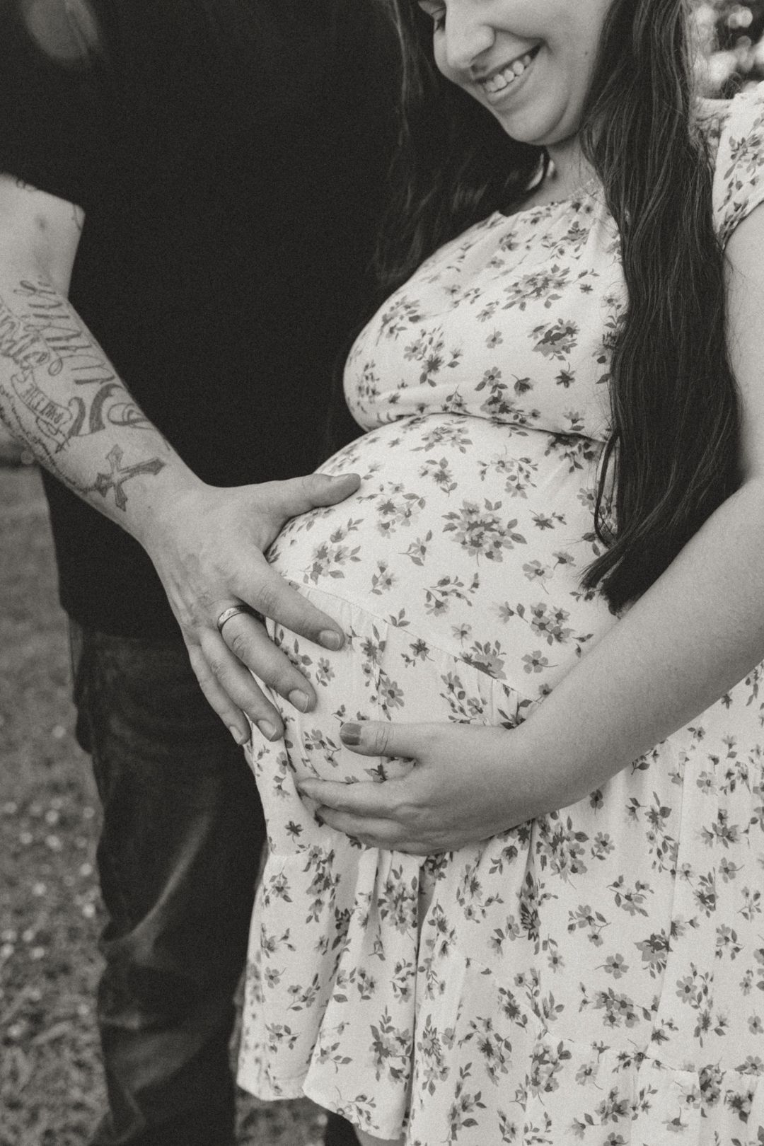 Black and white photo of pregnant woman in floral dress with tattooed partner cradling her baby bump.
