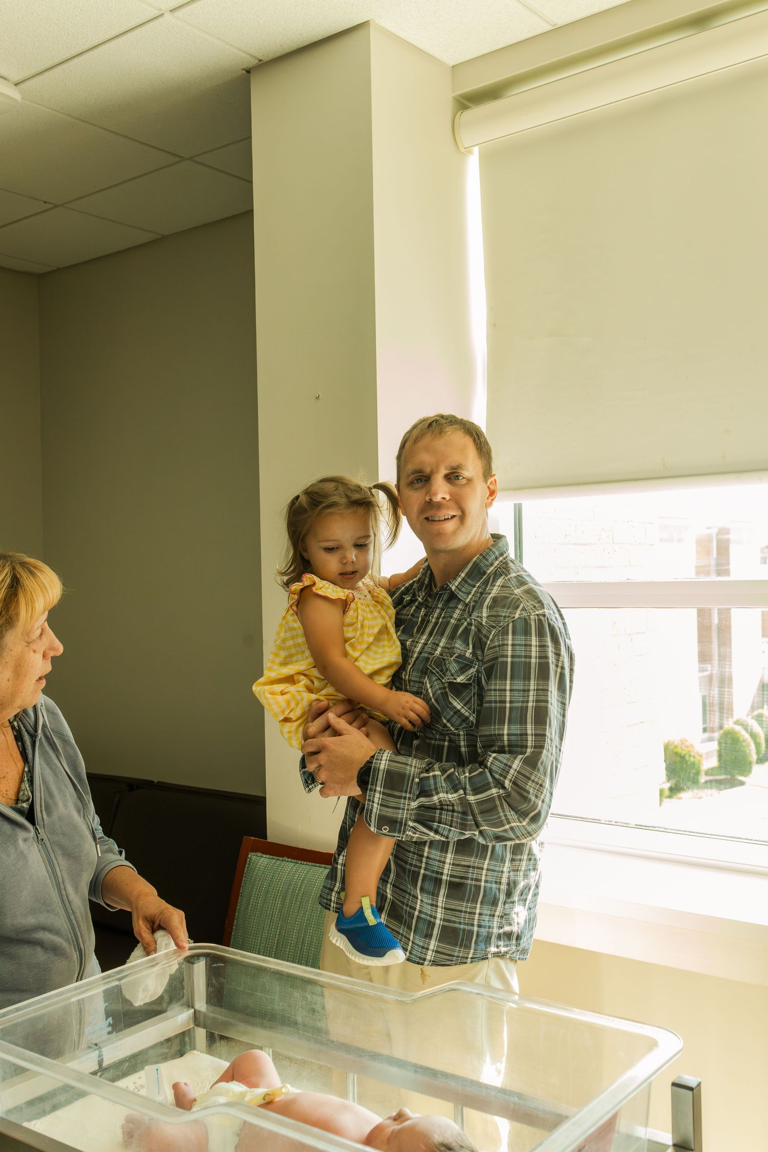 Smiling father holds toddler daughter beside newborn baby in hospital bassinet.