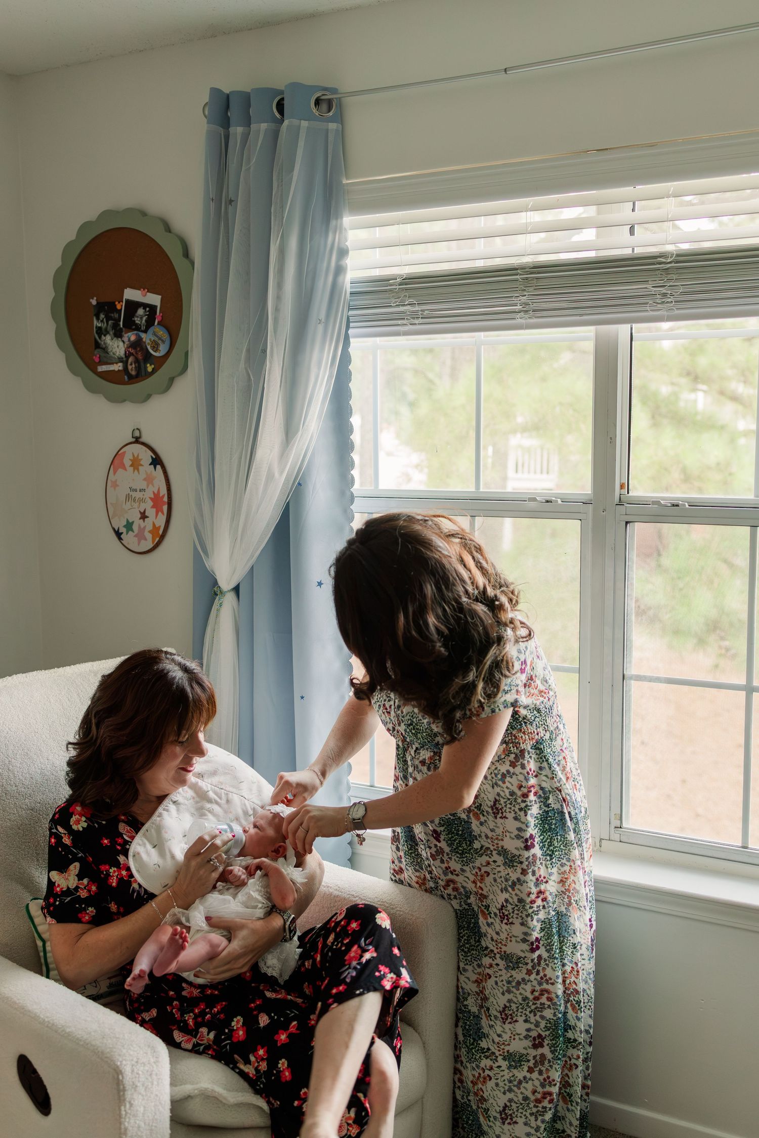 A mother sits in a nursery chair holding her newborn while an older girl in a floral dress leans in to look.
