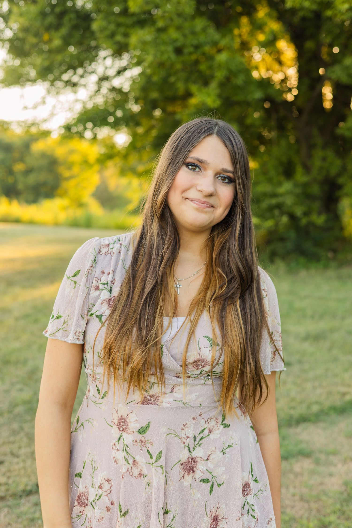A young woman with long brown hair wearing a floral dress smiling outdoors in golden hour light.