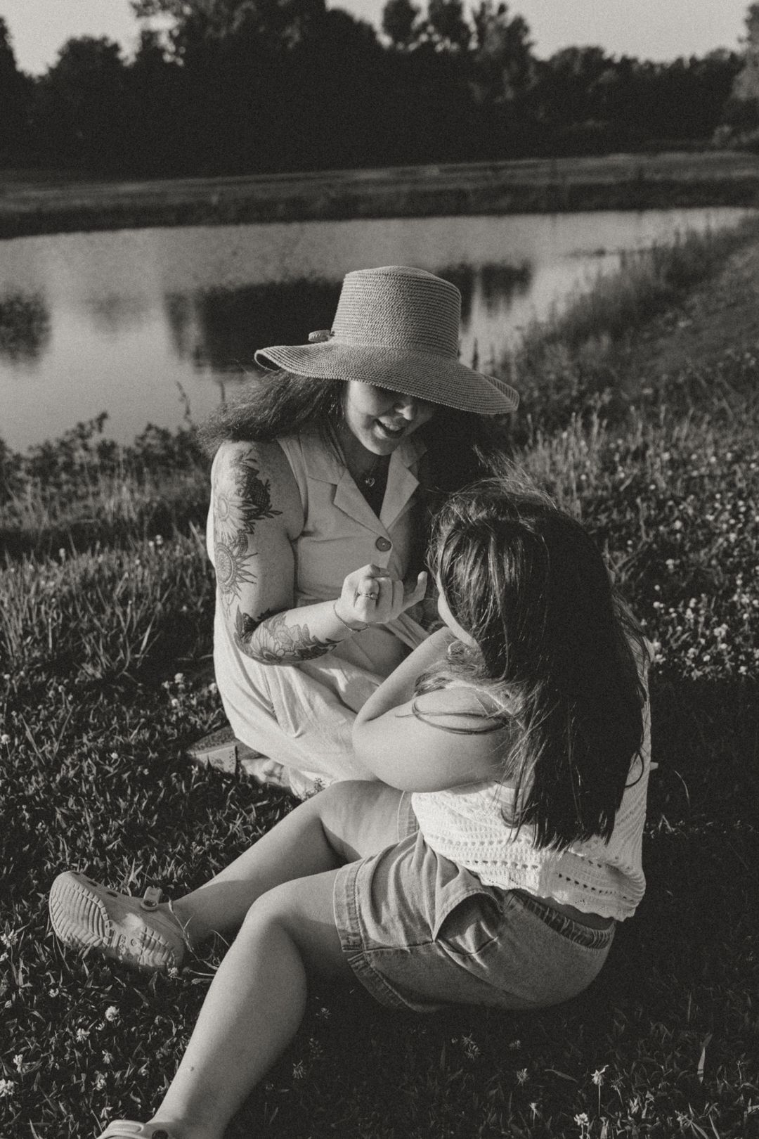 Black and white photo of a mother in a sun hat sitting on grass, lovingly holding her young daughter near a pond.
