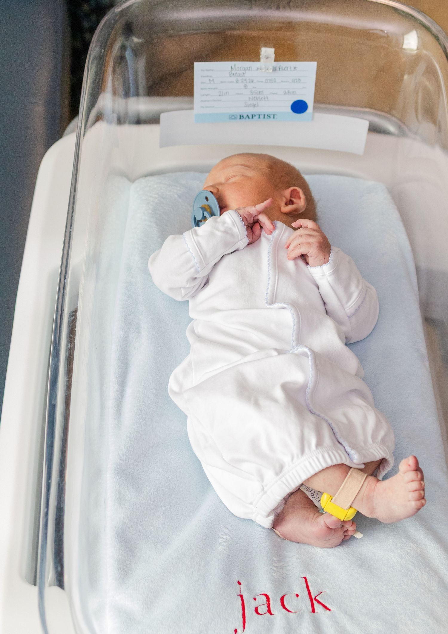 Newborn baby named Jack in white outfit resting in hospital bassinet with pacifier.