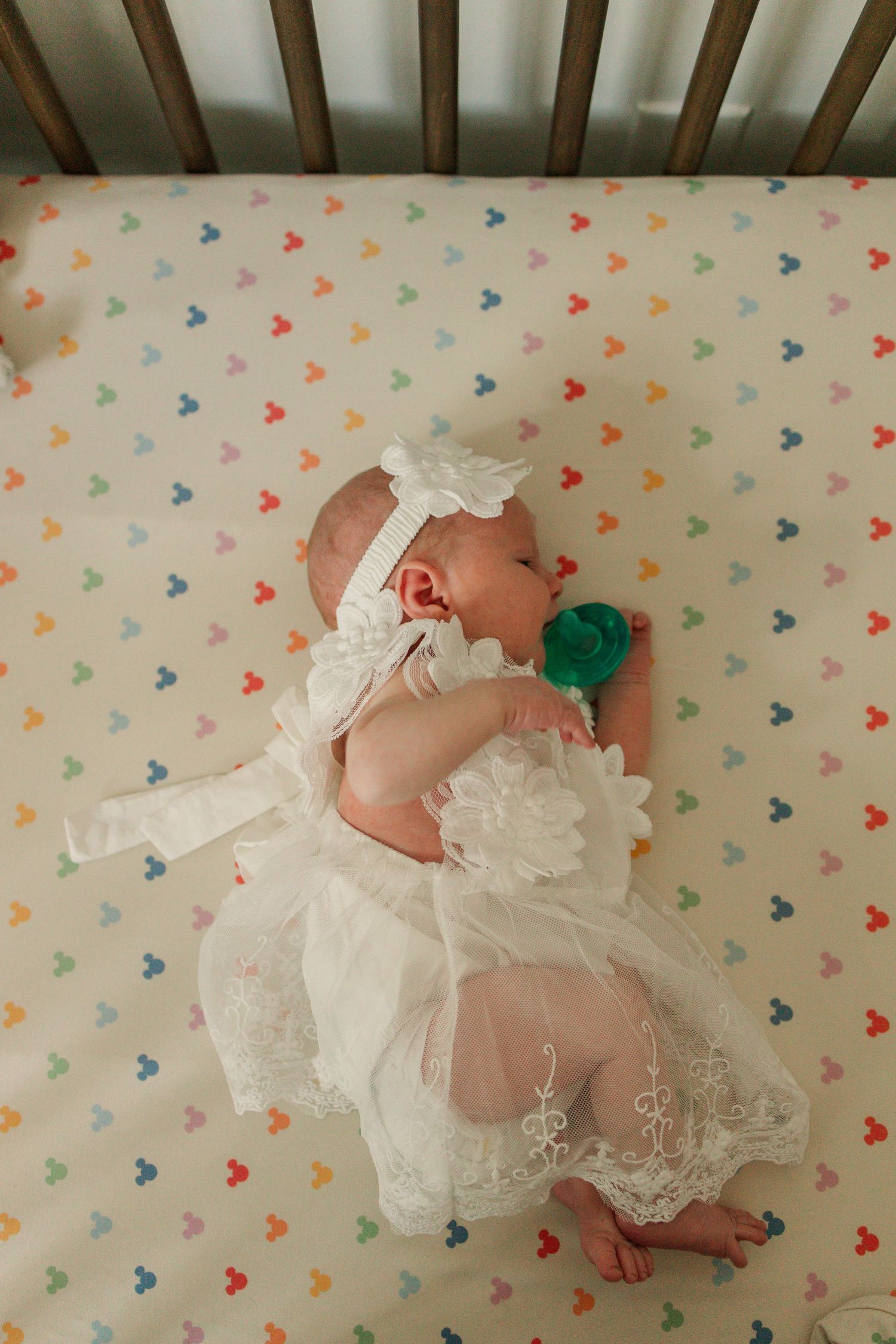 Newborn baby in white lace dress lying on colorful Mickey Mouse patterned crib sheet.