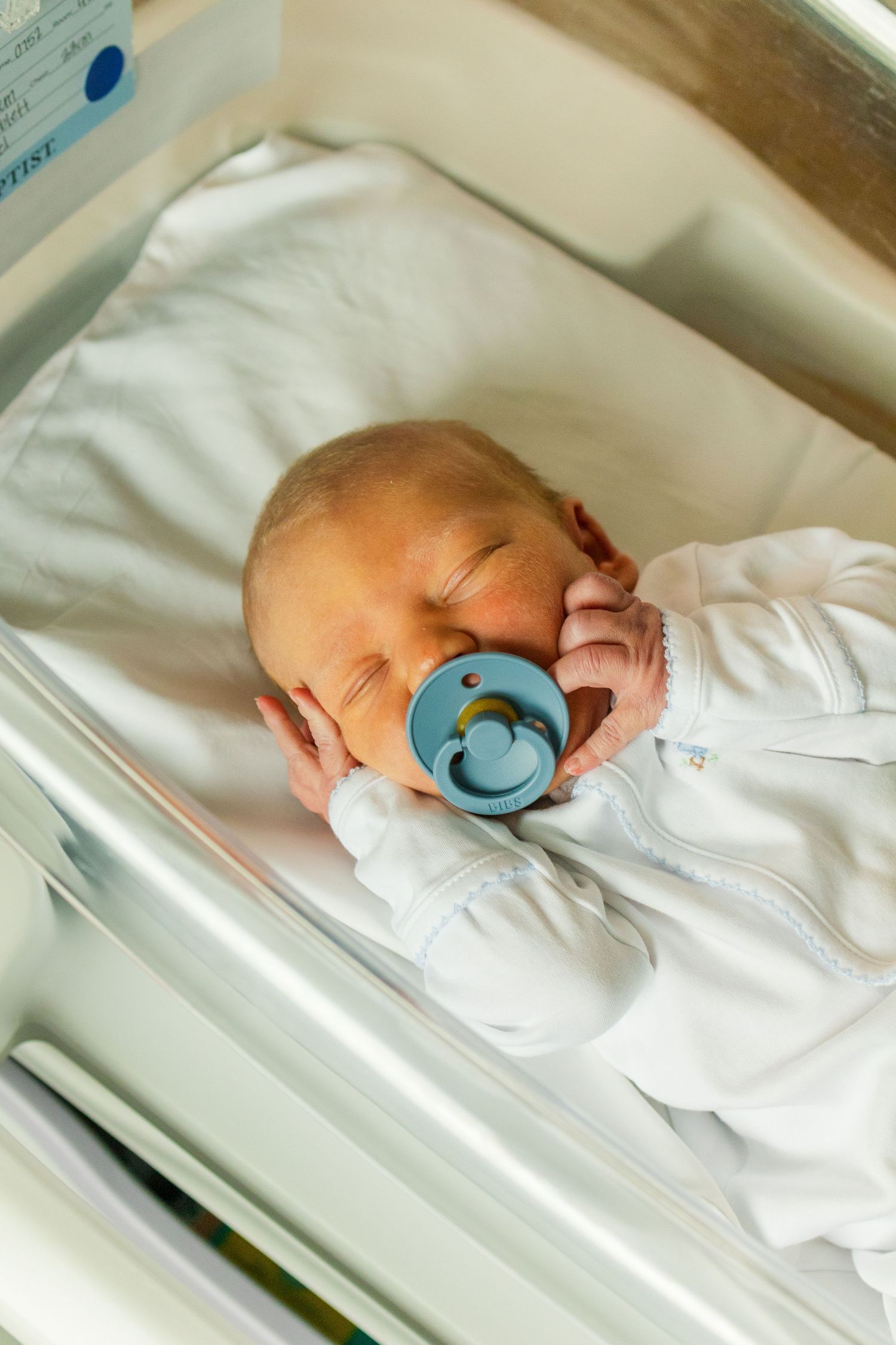 Peaceful newborn baby sleeping in hospital bassinet with blue pacifier in white outfit.