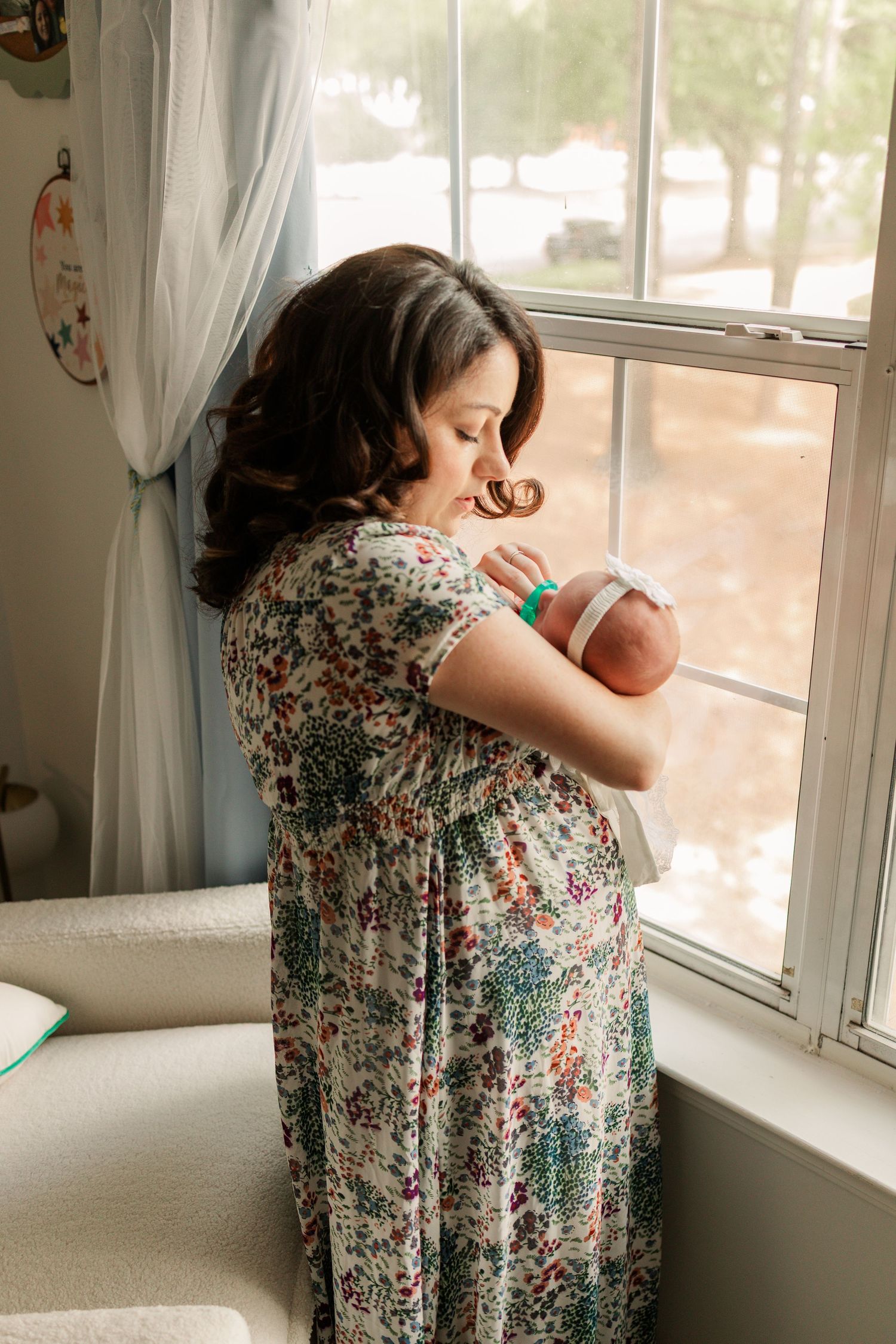 Mother in floral dress tenderly kisses her newborn baby while standing by a sunlit window.