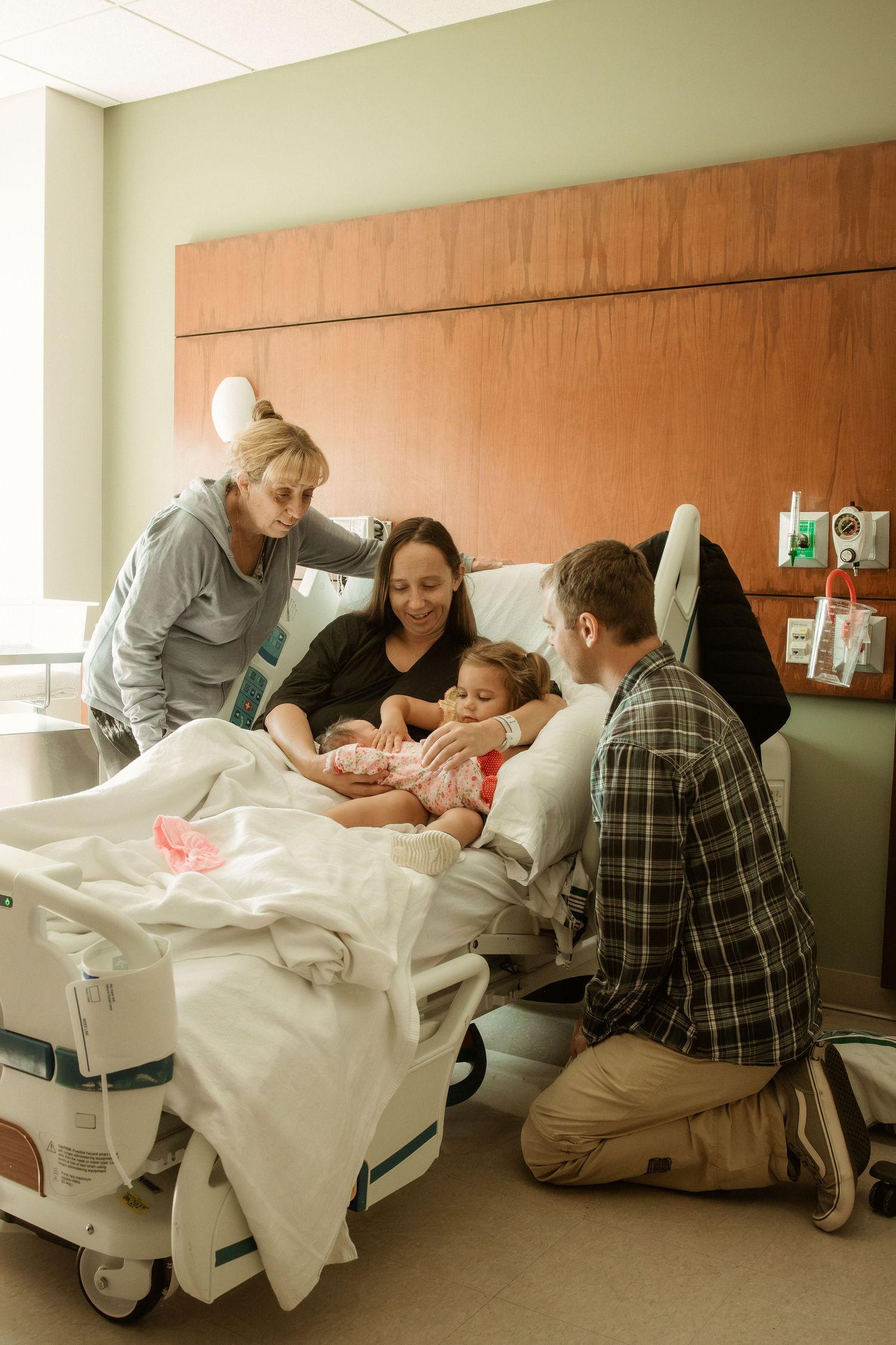 Family surrounds new mother holding newborn and toddler in hospital room.
