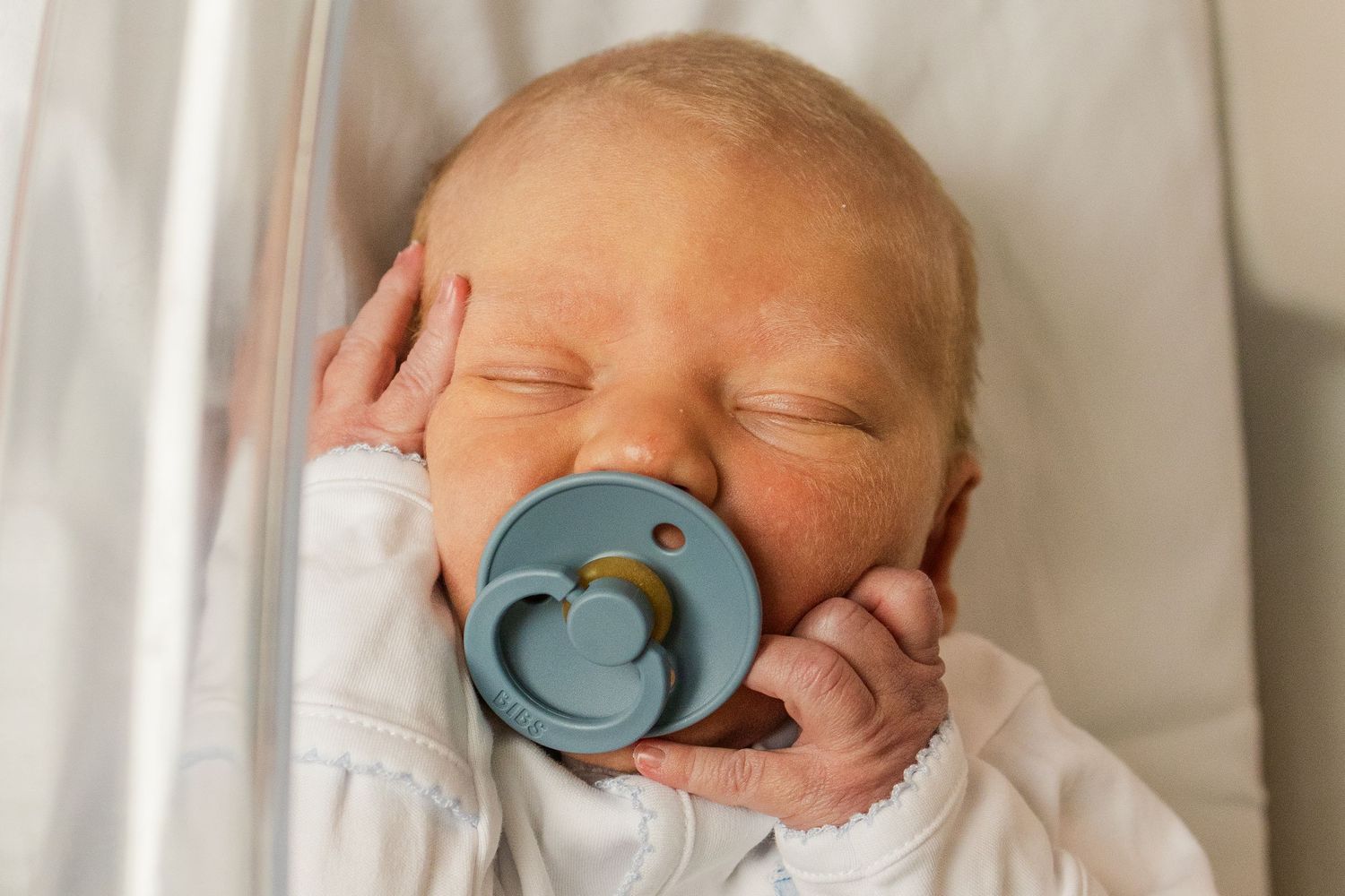 Peaceful newborn baby sleeping with a blue pacifier, wrapped in white hospital blanket.