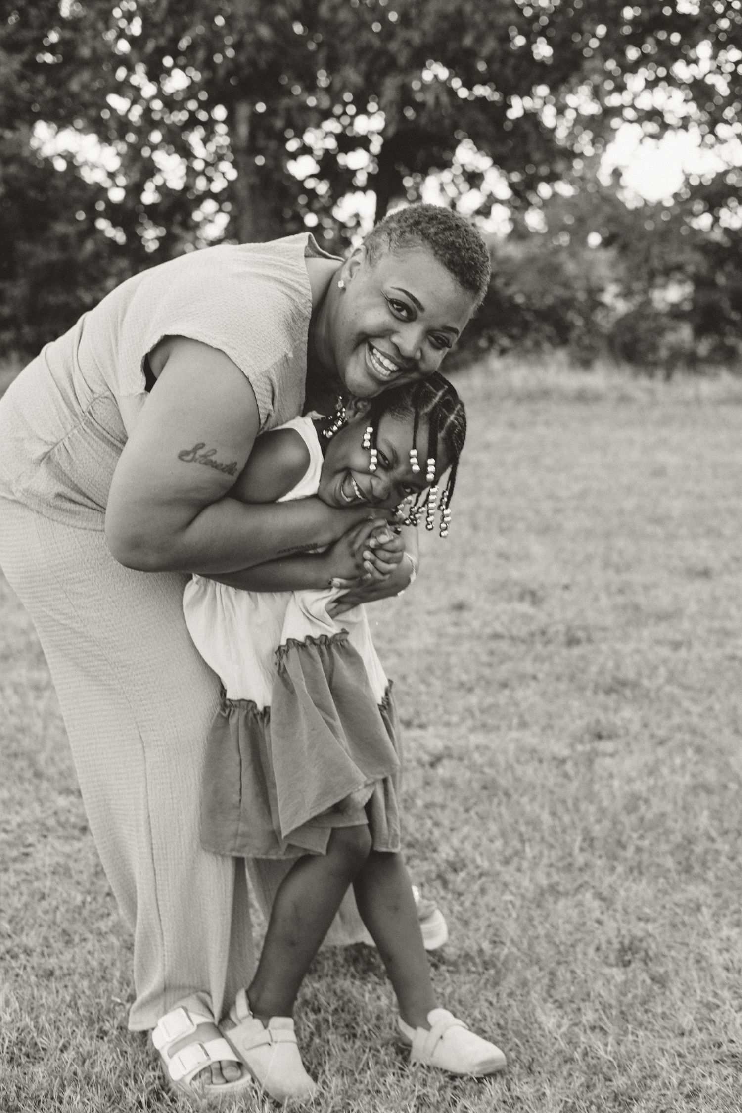 Black and white photo of a smiling woman hugging a young girl with braids outdoors on green grass.