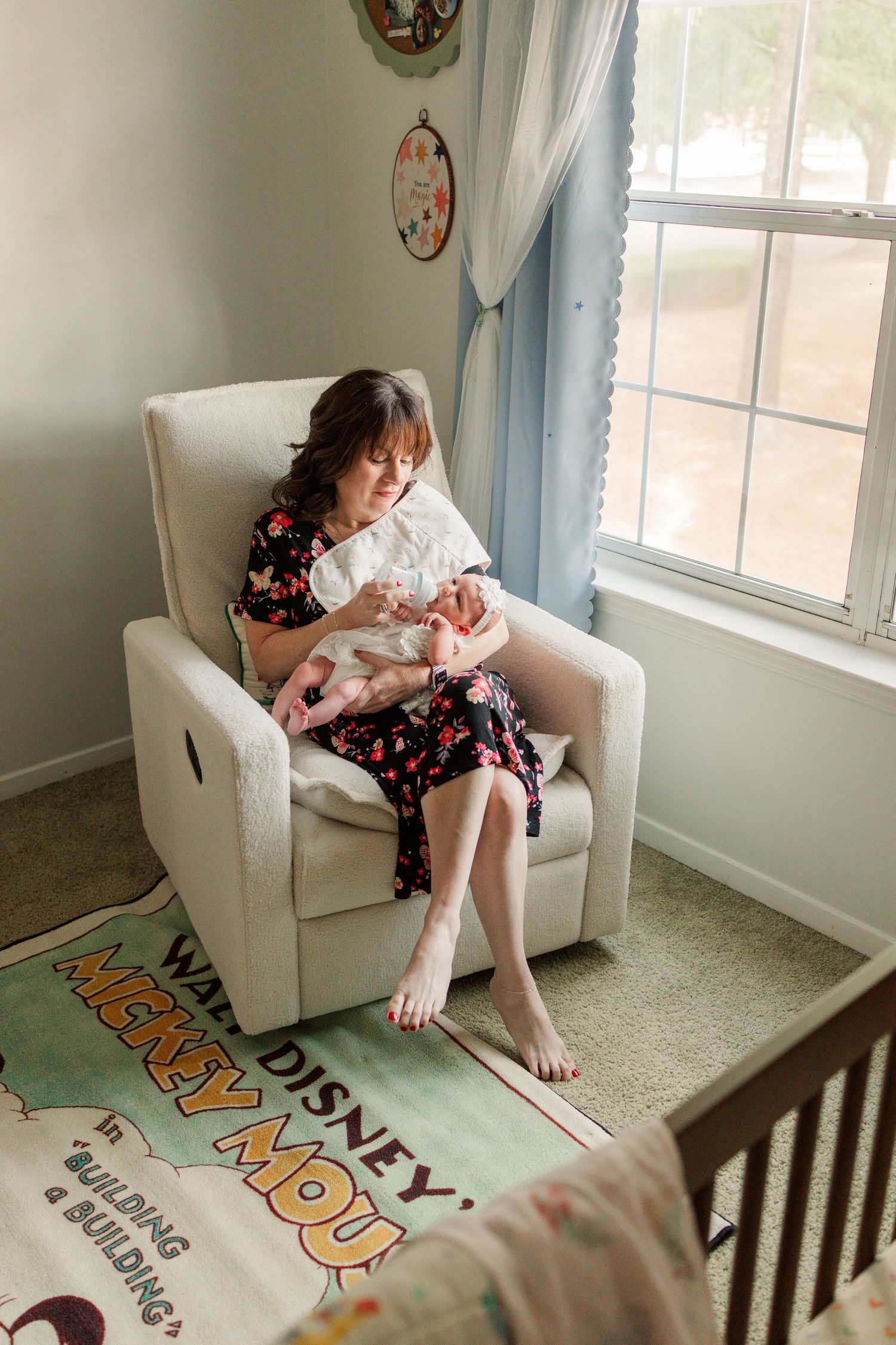 Mother in floral dress sits in nursery glider chair cradling her sleeping newborn by window.