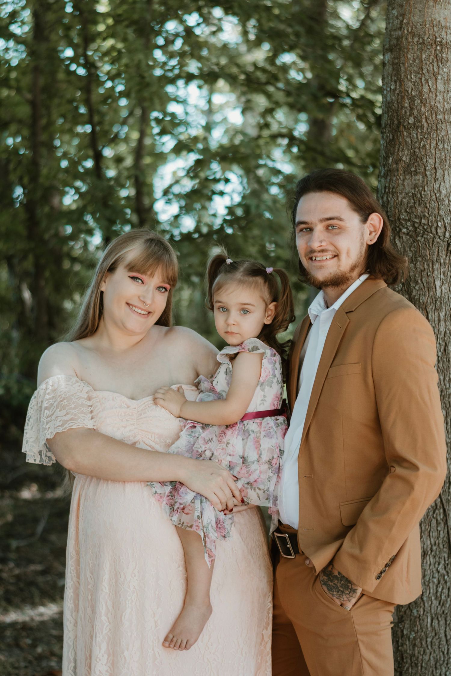 Smiling pregnant woman in pink lace dress and man in tan suit hold young girl in floral dress outdoors.