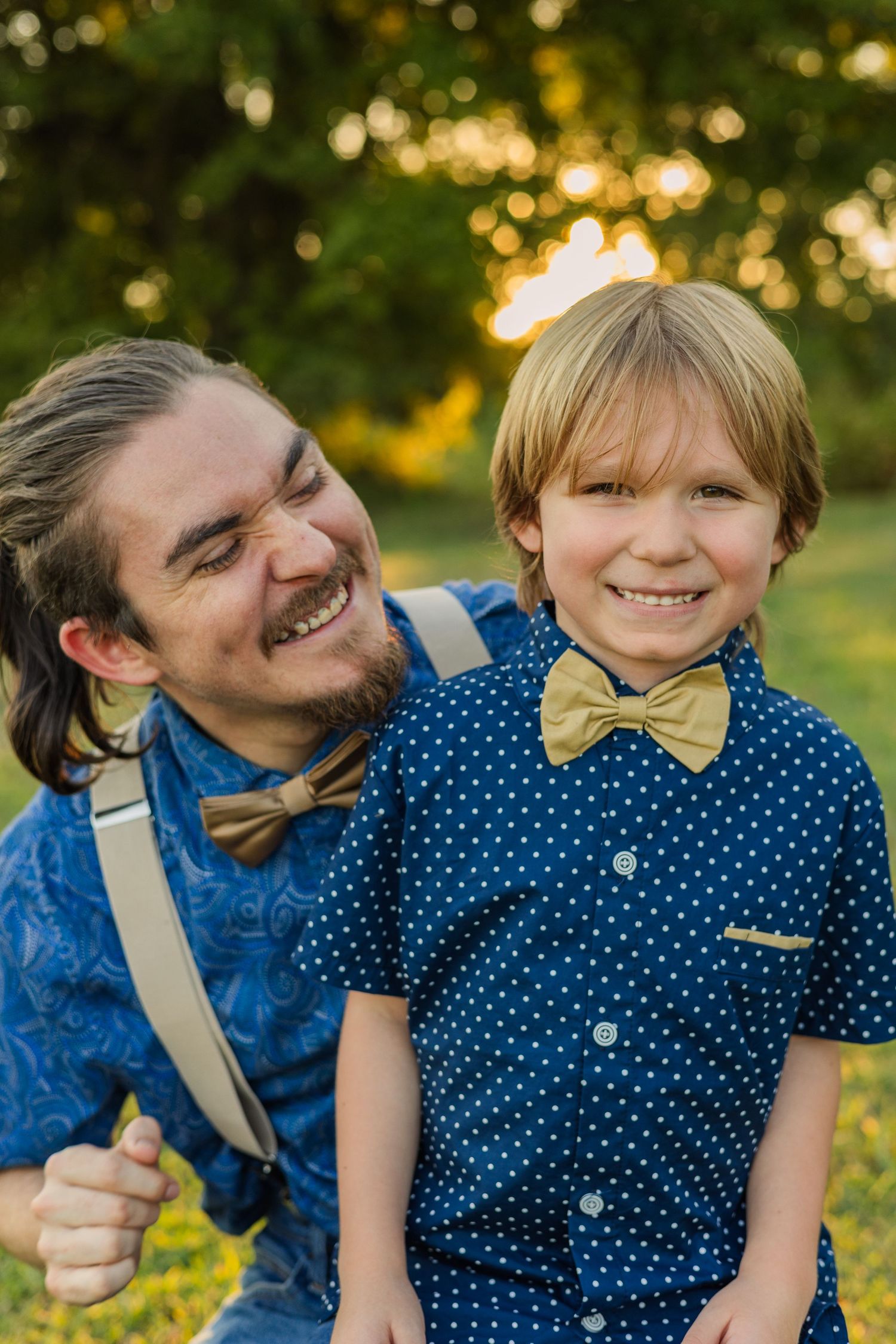 Laughing father crouches beside his young son, both wearing matching blue shirts and gold bow ties outdoors.