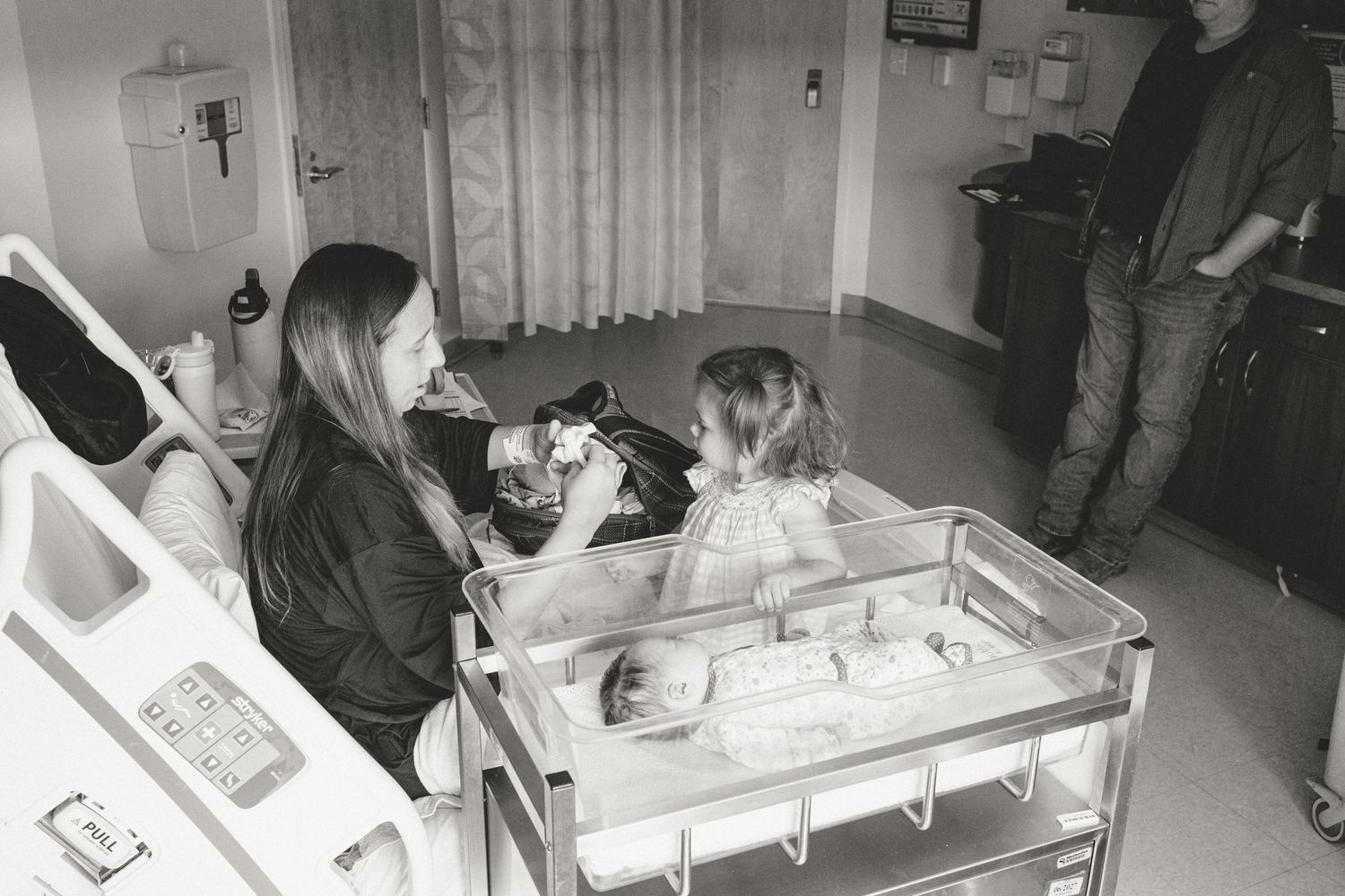 Black and white photo of mother and daughter with newborn in hospital bassinet.