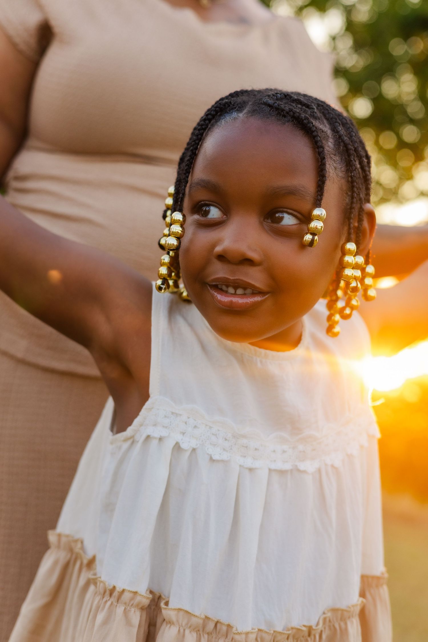 A young Black girl with gold beaded braids in a white dress smiles outdoors at golden hour sunset.