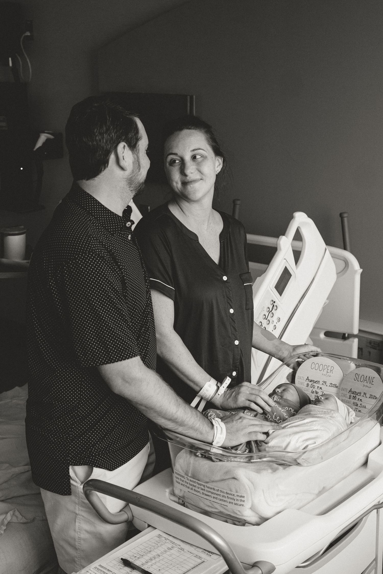 Black and white photo of new parents smiling at each other beside their newborn in a hospital bassinet.