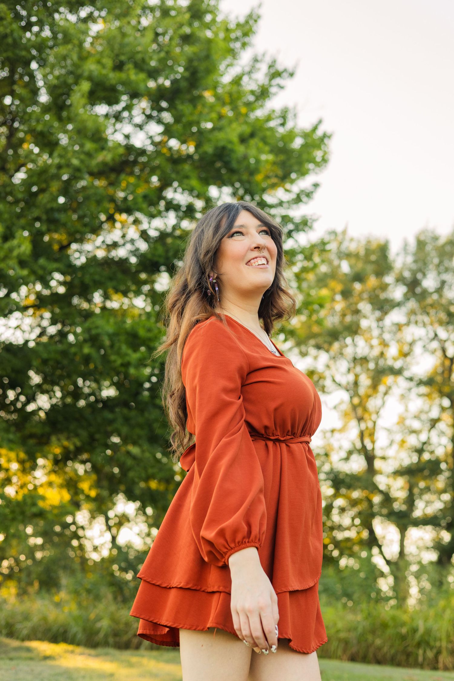 Woman in rust orange dress smiling and looking up while standing outdoors surrounded by green trees.