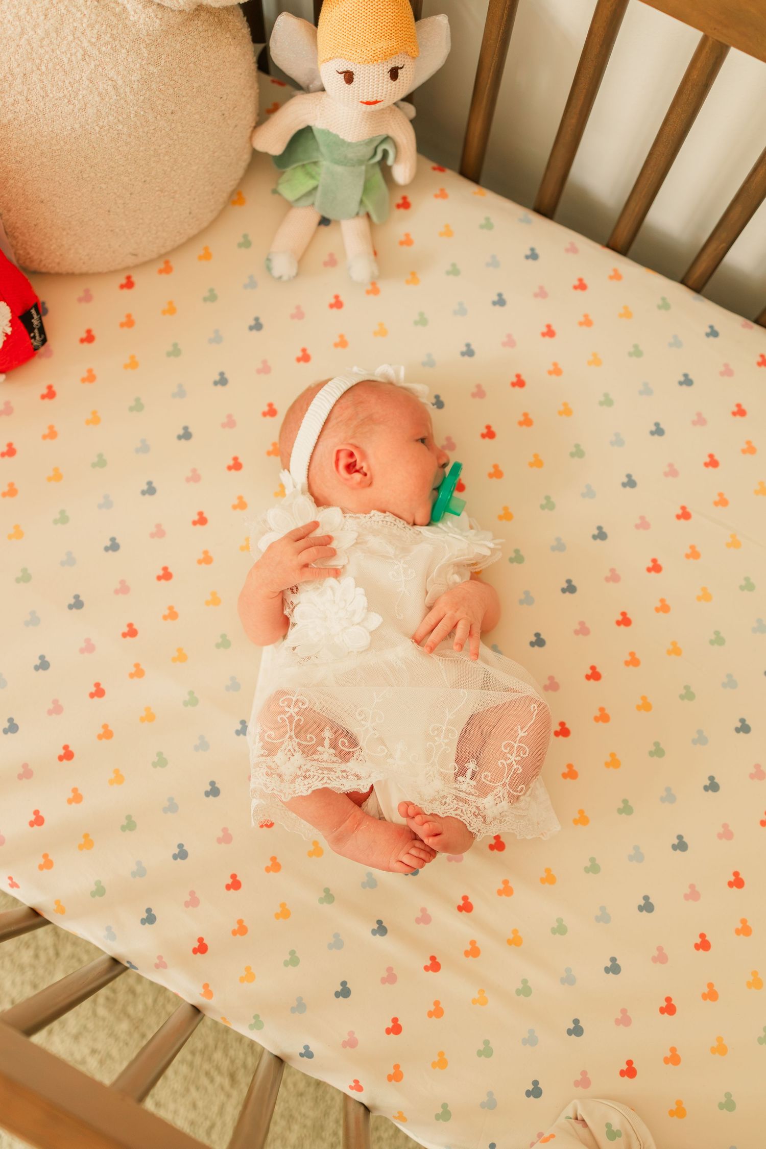 Newborn baby in white lace dress with pacifier sleeping in wooden crib with Disney sheet.