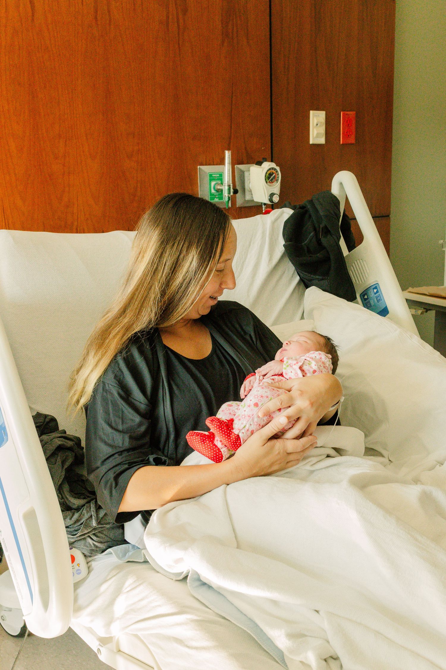 New mother sitting in hospital bed holding her newborn baby, wearing a black shirt, smiling lovingly at her infant.