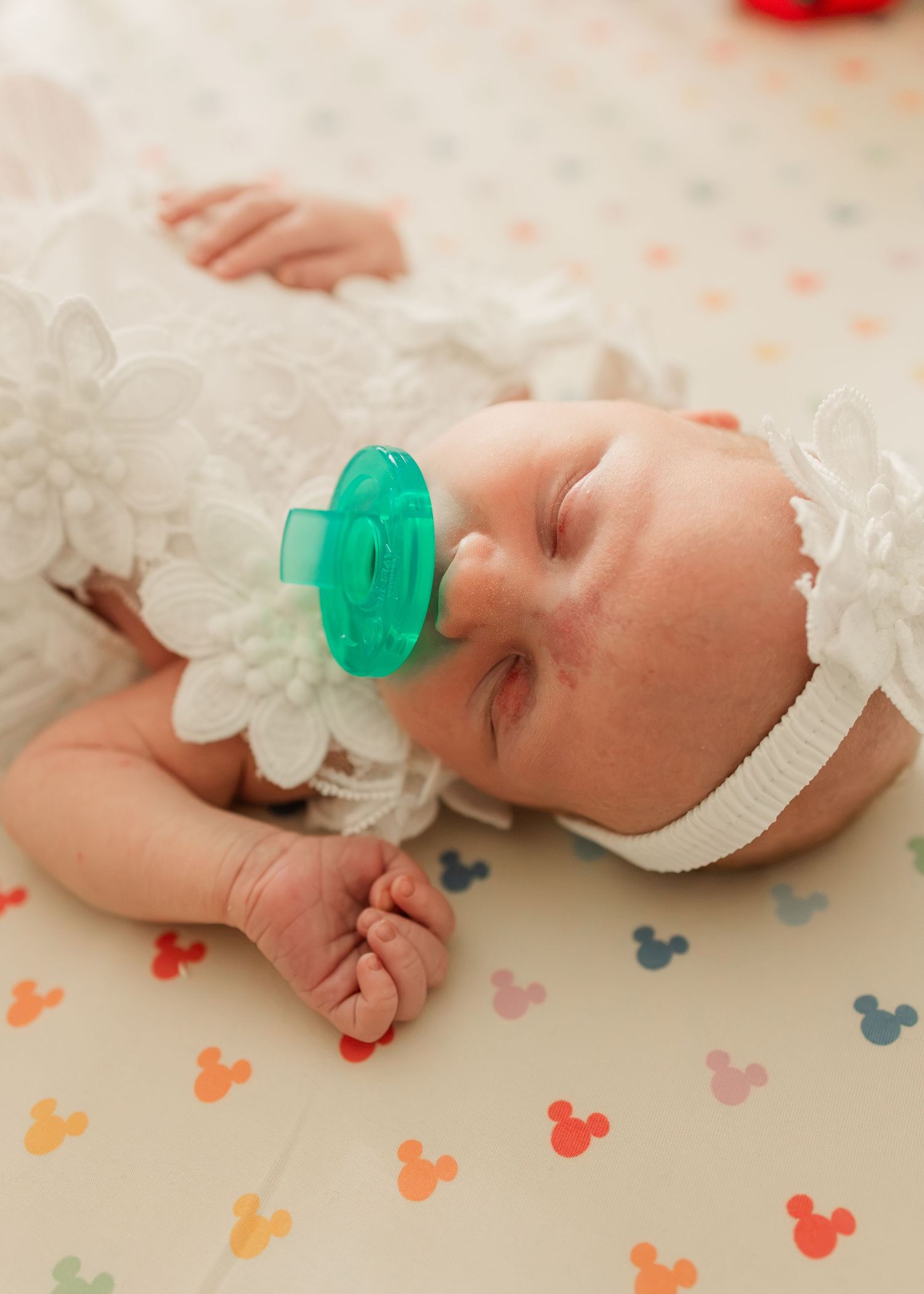 Close-up of peaceful newborn in white lace outfit with floral headband and teal pacifier sleeping.