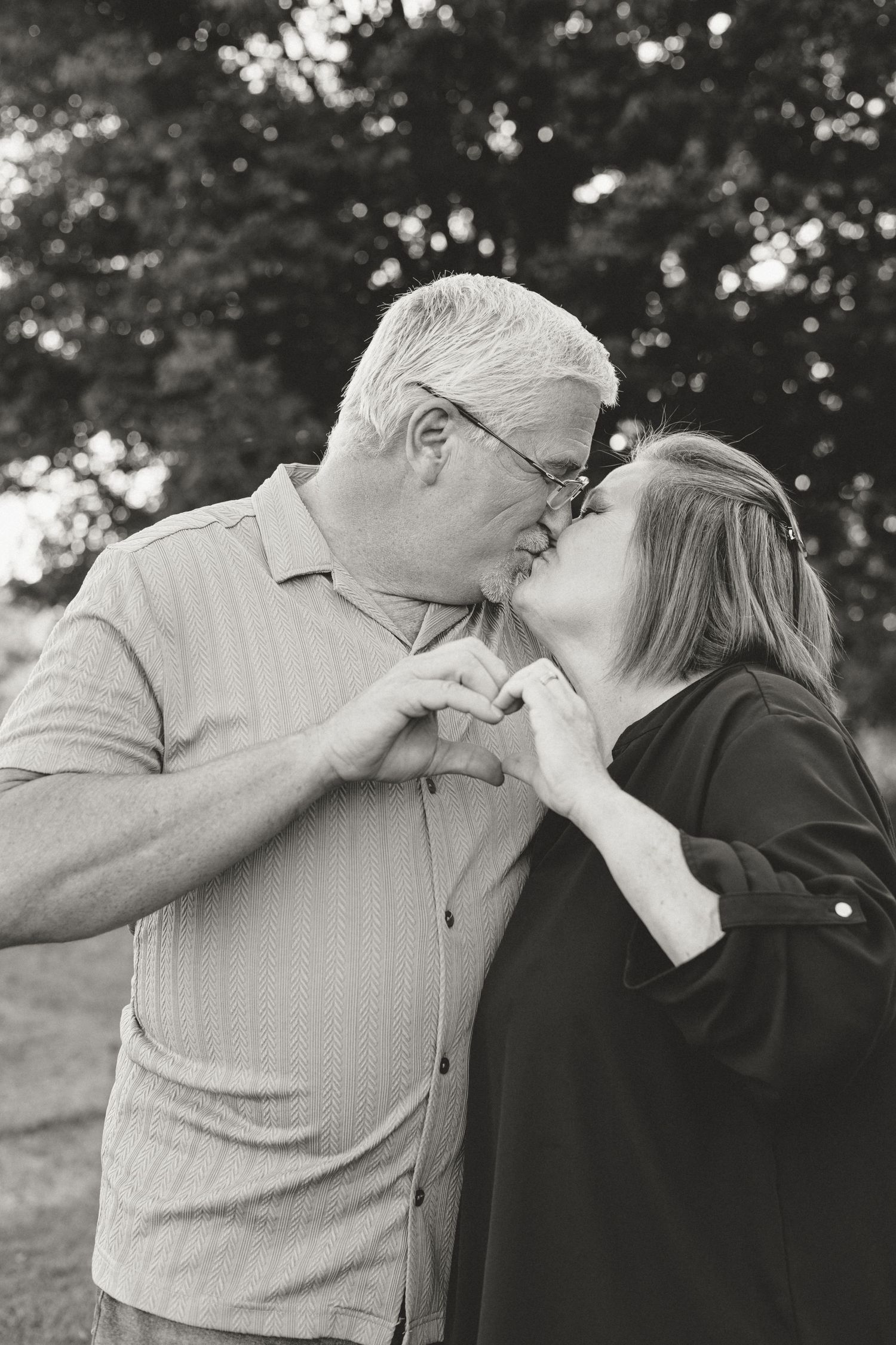 Older couple sharing a kiss while making a heart shape with their hands in a black and white outdoor portrait.