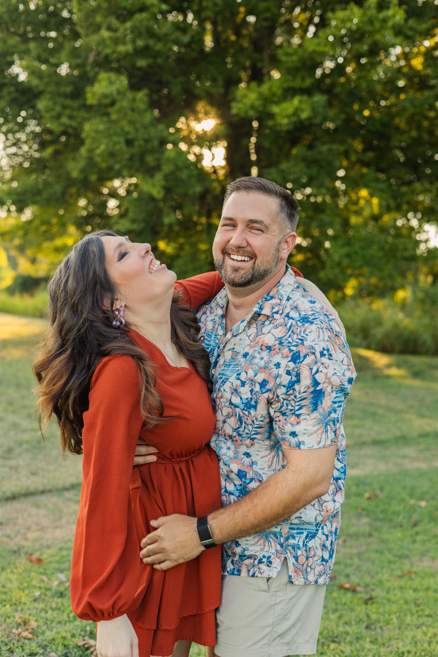 Laughing couple embracing outdoors during golden hour, woman in red dress and man in floral shirt.
