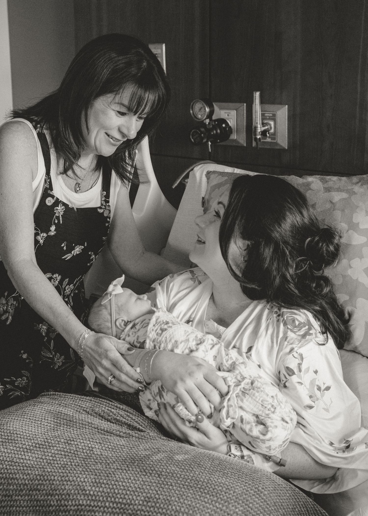 Black and white photo of two women tenderly holding a newborn baby while sitting on a bed.