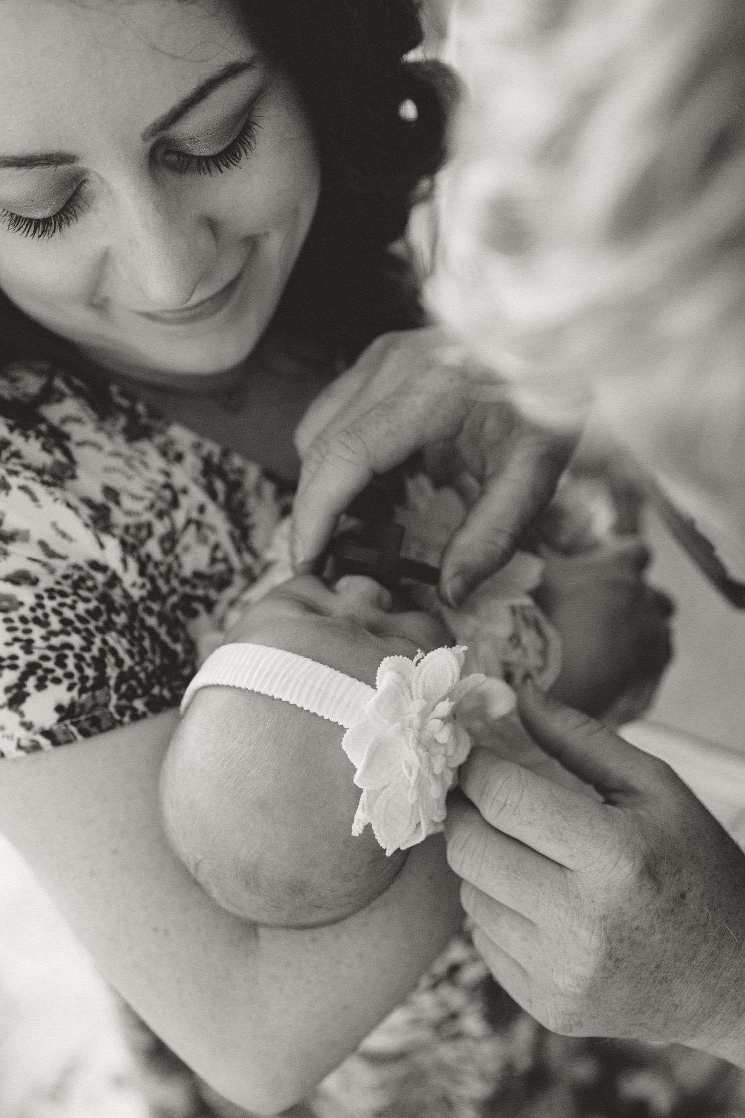 Black and white image of mother smiling as someone adjusts white floral headband on newborn.