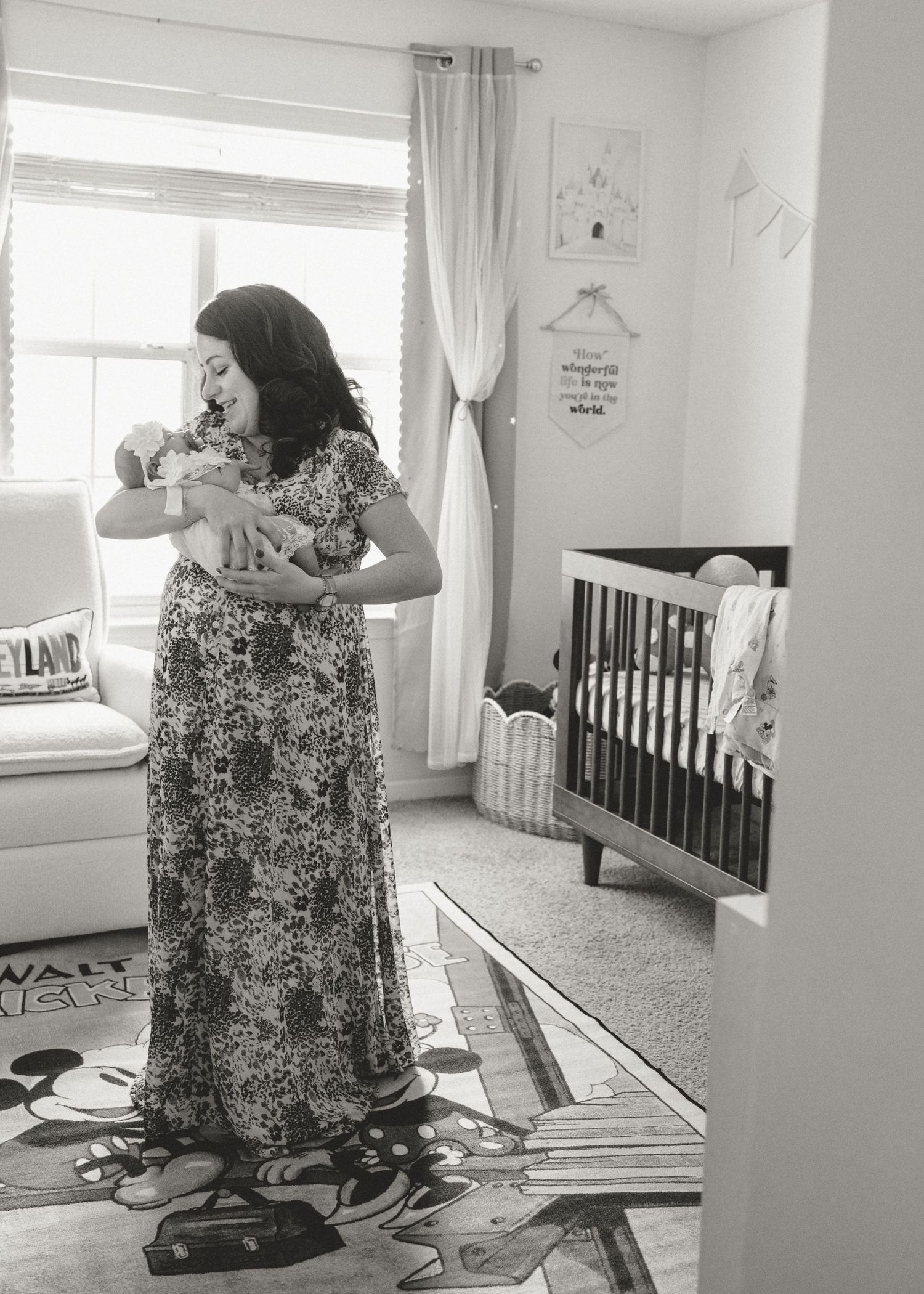 Black and white photo of a pregnant woman in floral dress holding a newborn in a nursery with a crib.