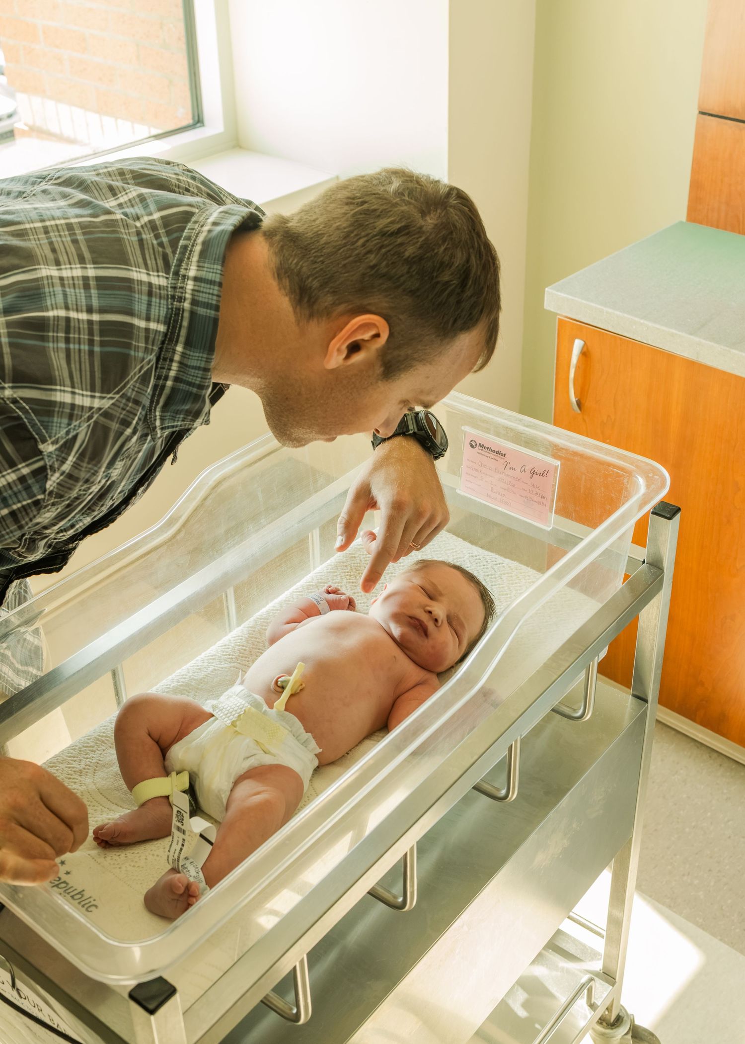 Father leaning over hospital bassinet touching newborn baby in maternity ward.