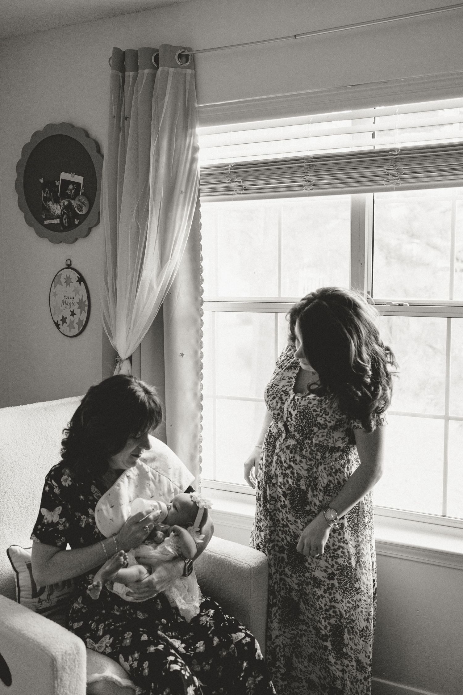 Black and white photo of a grandmother holding a newborn while a woman in a floral dress stands nearby.