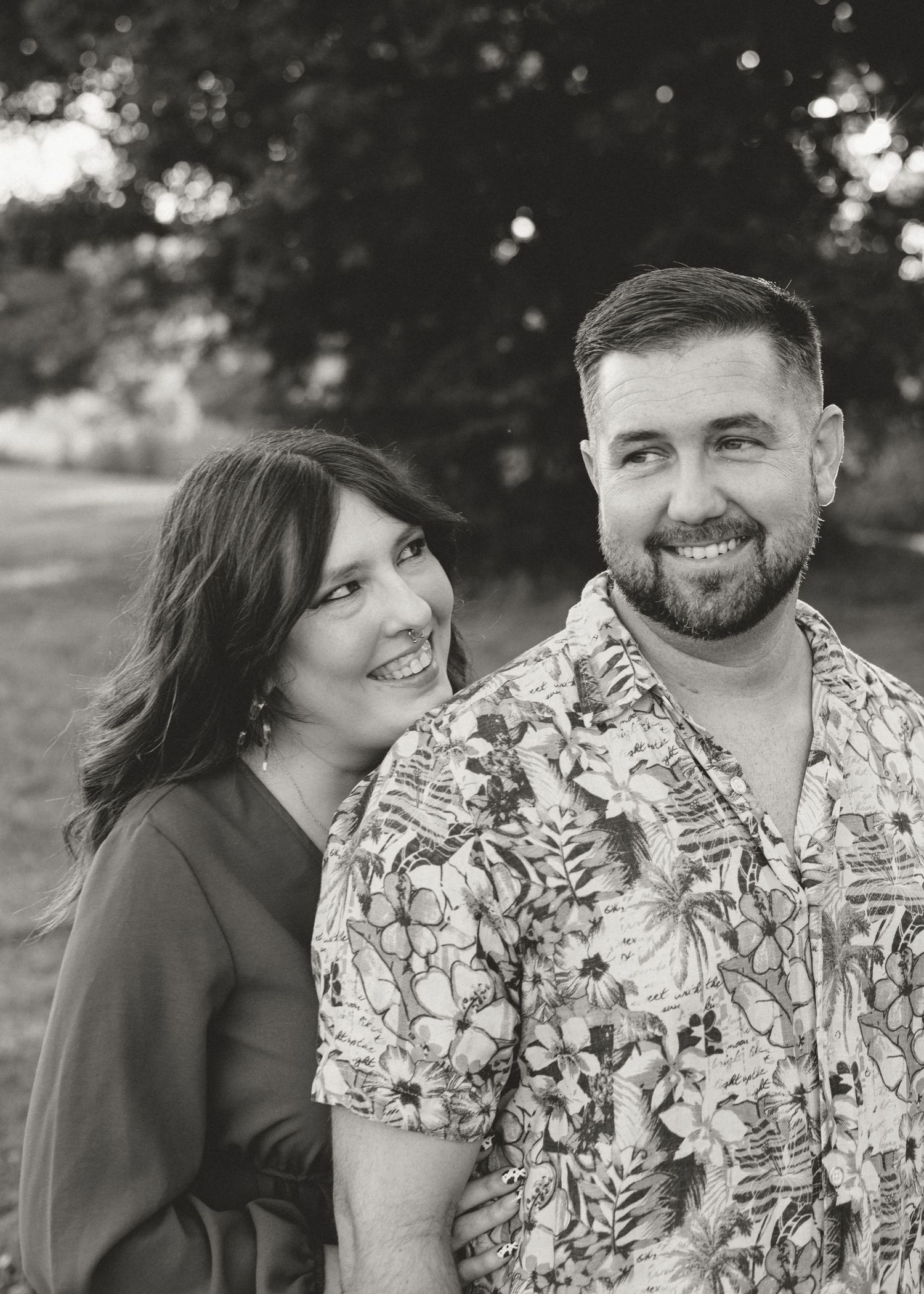 Black and white portrait of a smiling couple posing together outdoors among trees.