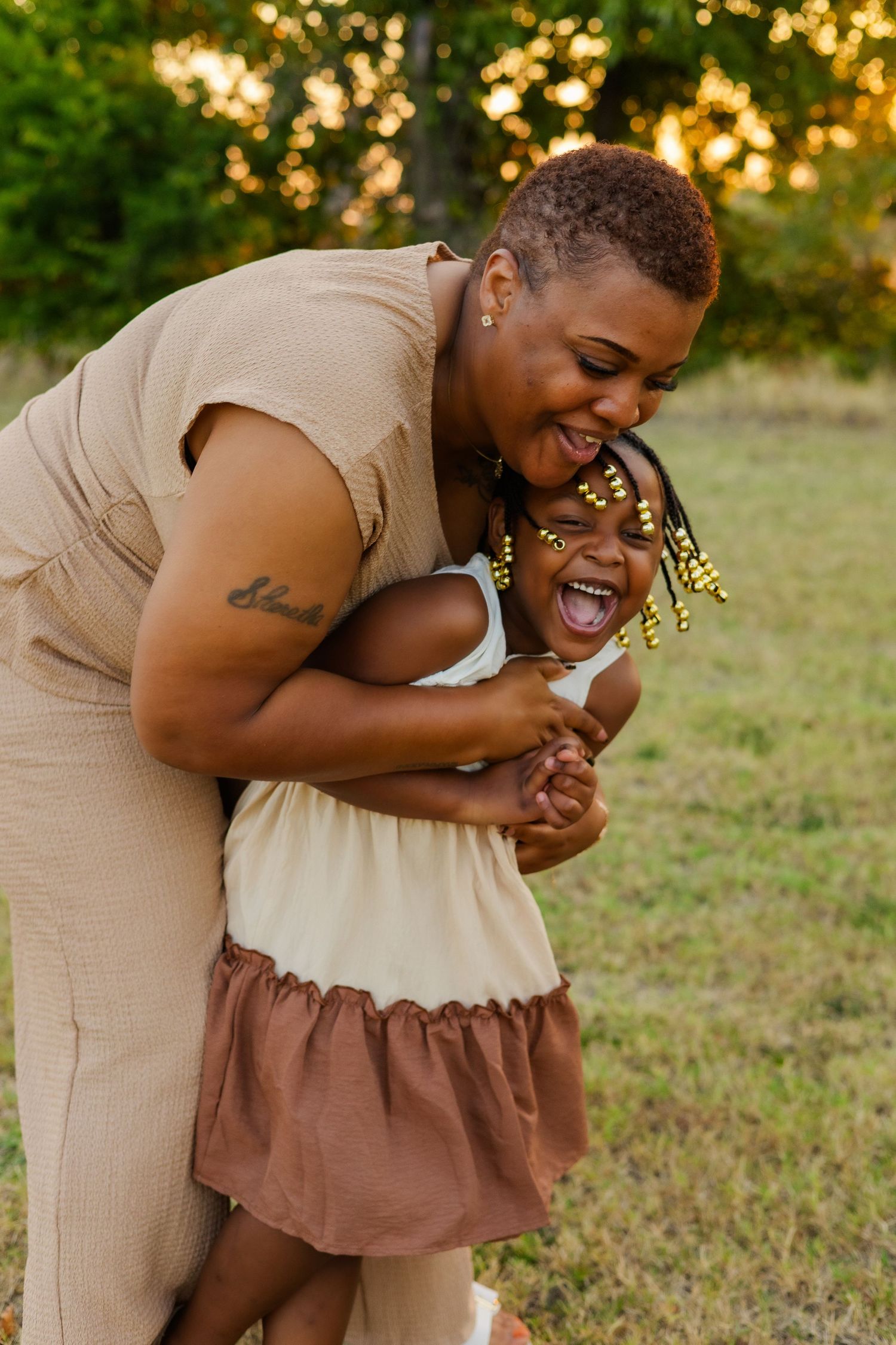 Joyful Black mother hugs laughing little girl with beaded braids in a sunny green park.