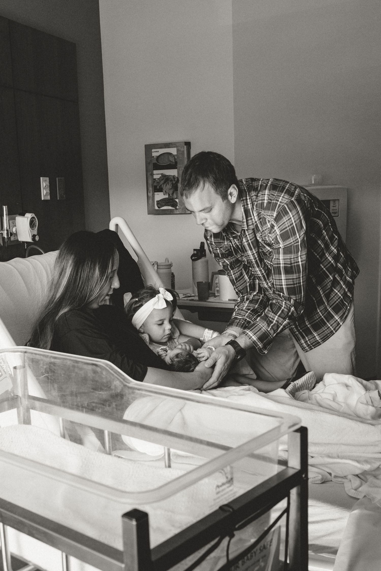 Black and white photo of parents with toddler meeting newborn baby in hospital room.
