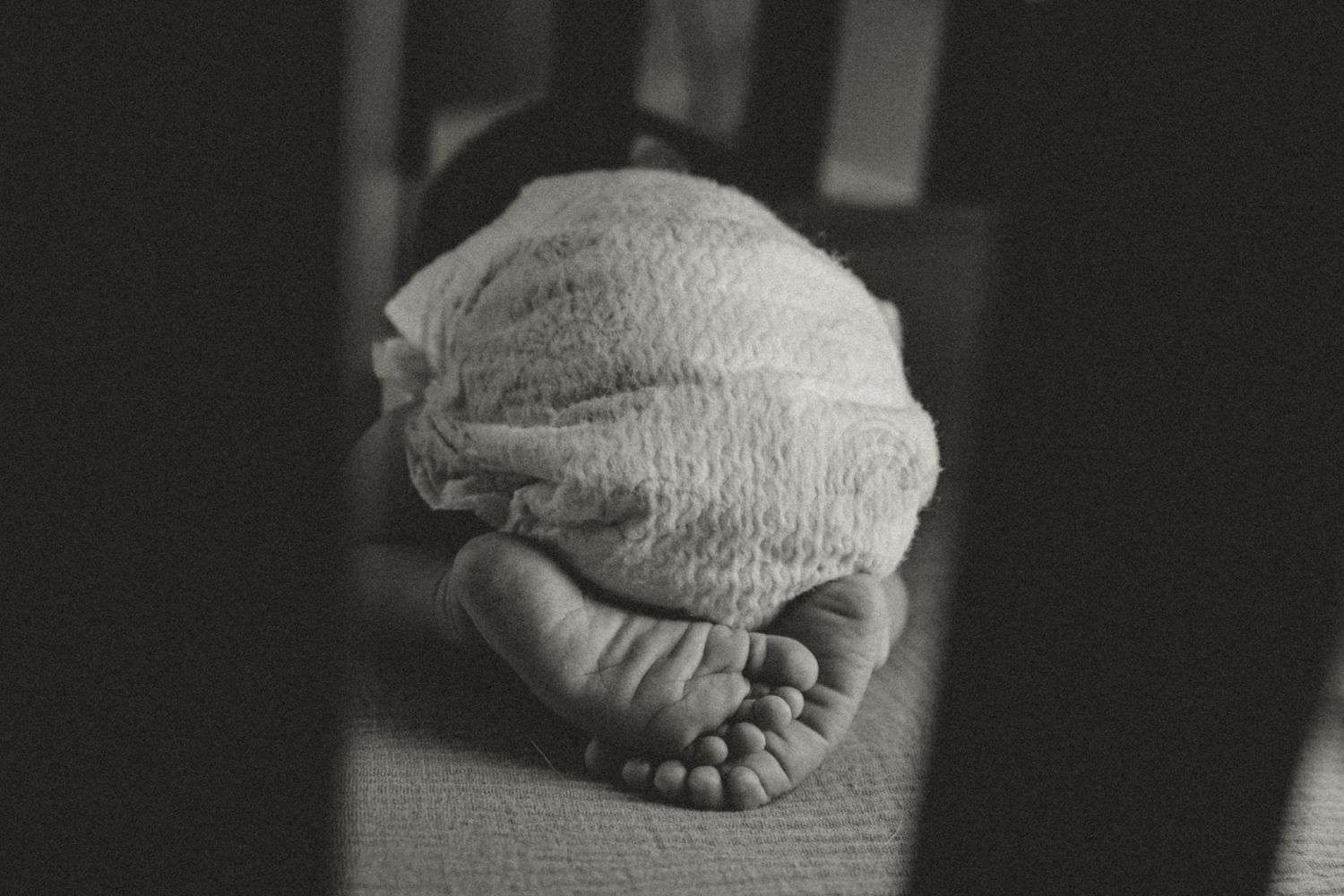 Black and white photo of a sleeping newborn baby wrapped in white swaddle blanket with tiny feet visible.