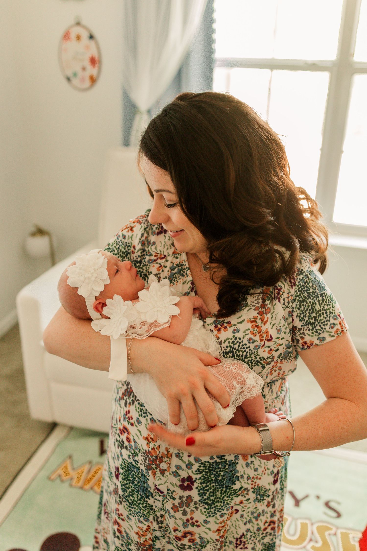 A mother in a floral dress tenderly holds her newborn baby wearing a white lace headband in a bright nursery.