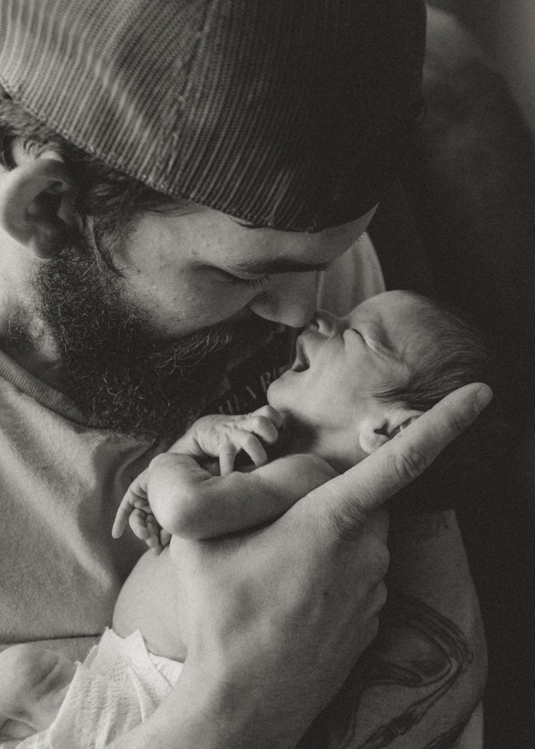 Bearded father in cap tenderly holds newborn baby close, sharing an intimate moment in black and white photo.