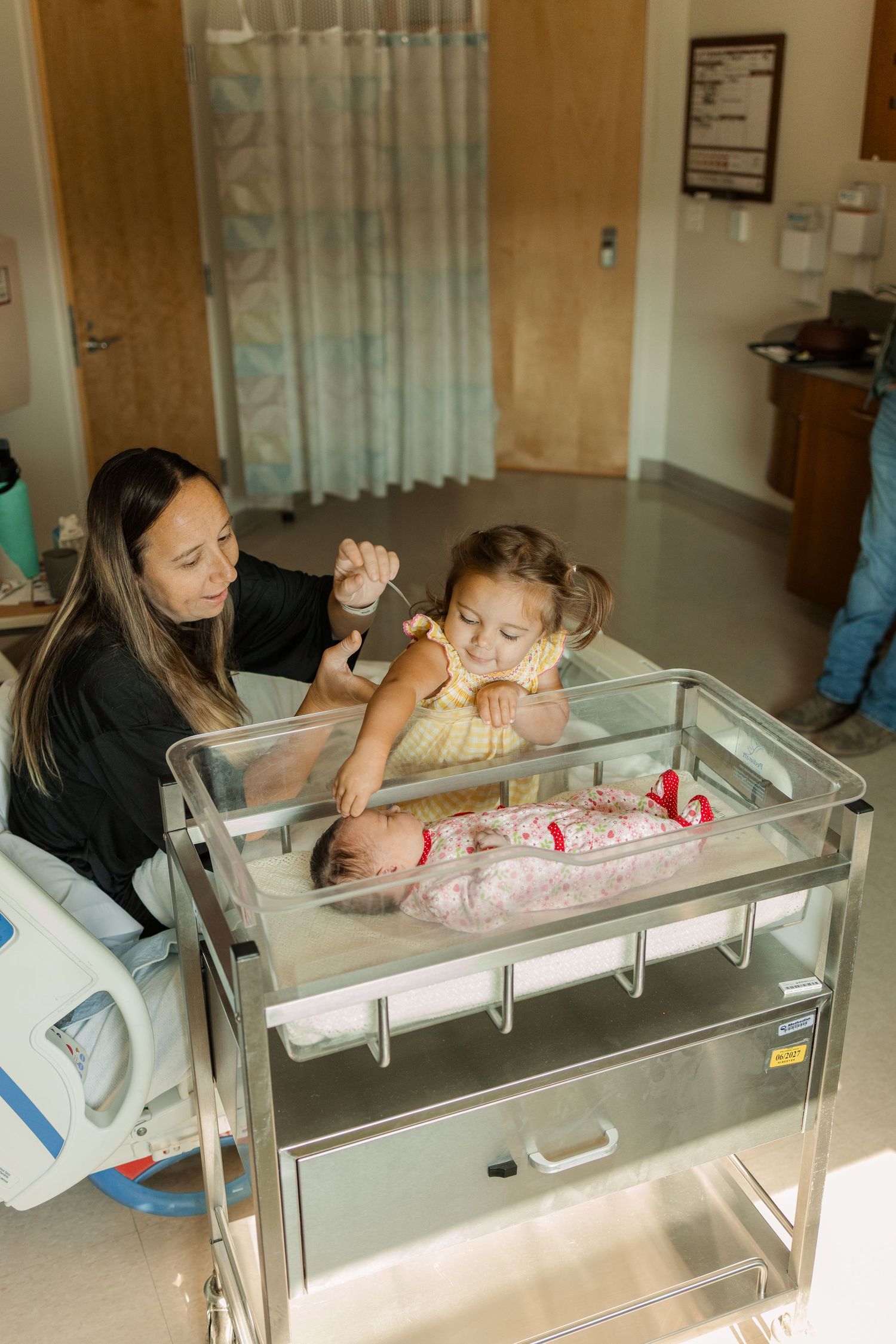 Mother and young girl looking at swaddled newborn in hospital bassinet.
