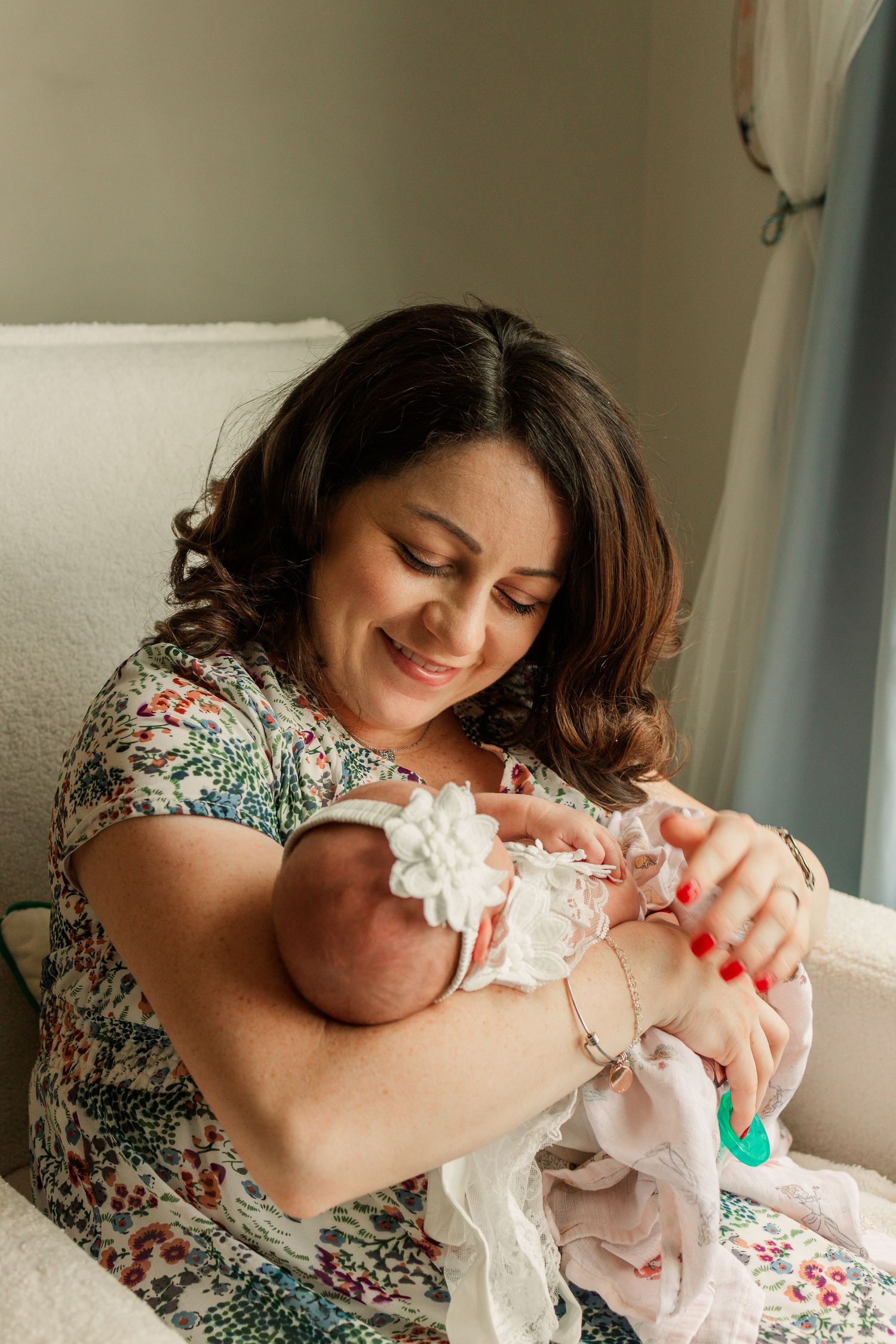 A smiling brunette mother in a floral dress tenderly holds her newborn baby with a white flower headband.