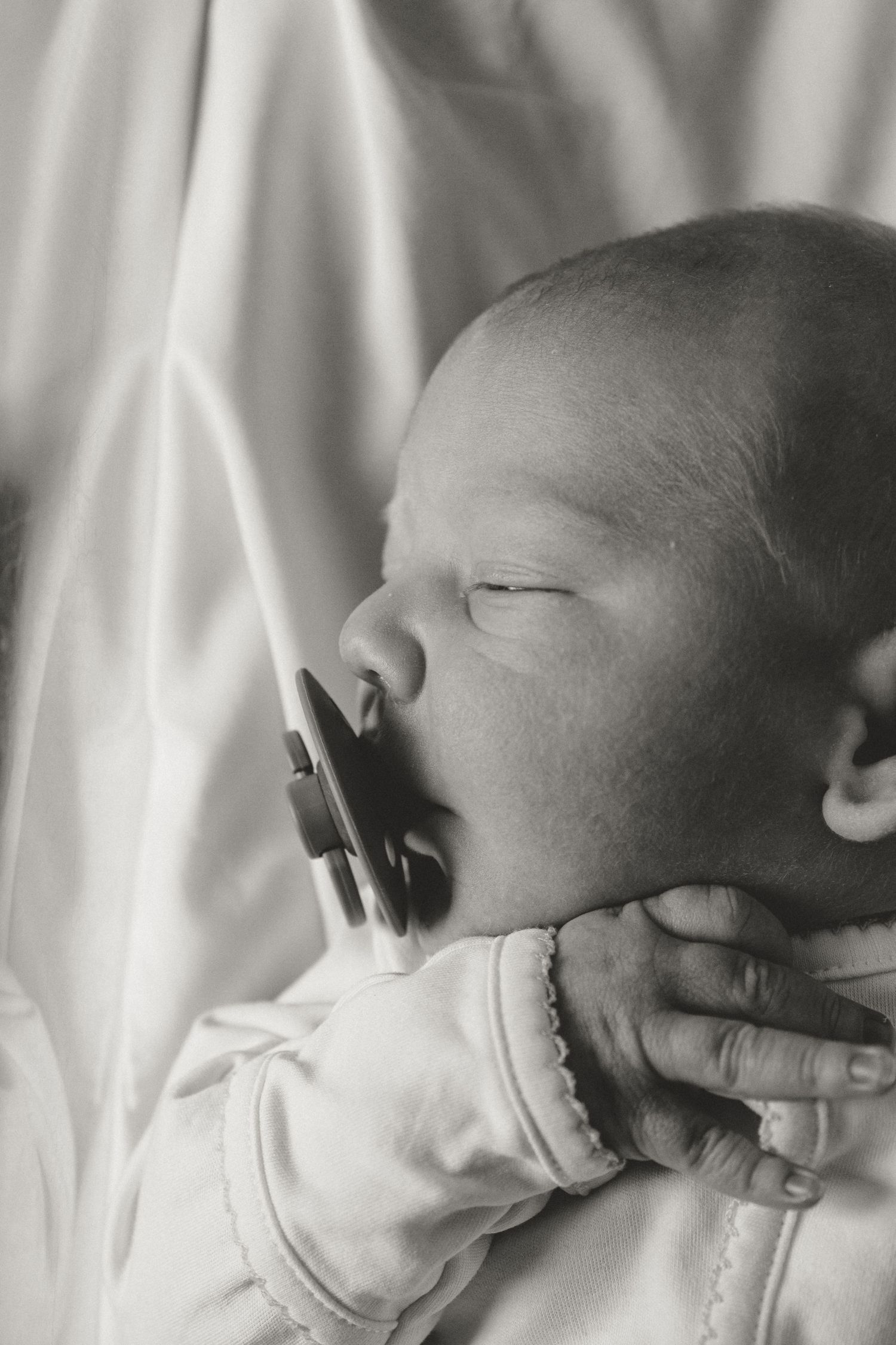 Black and white close-up of a sleeping newborn baby with a pacifier held gently by a parent's hand.