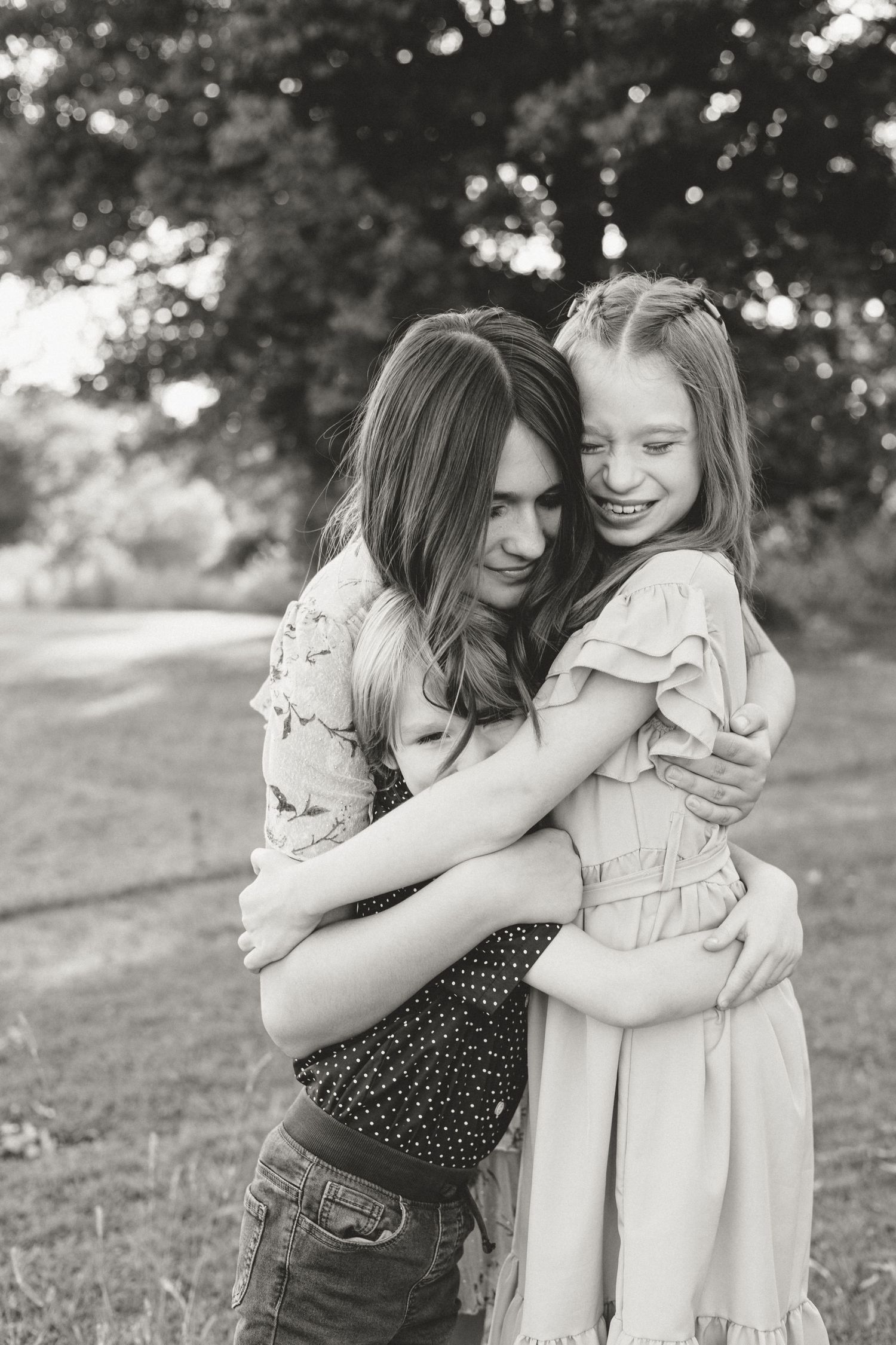 Black and white photo of two young girls and their brother hugging and laughing together in an outdoor setting.